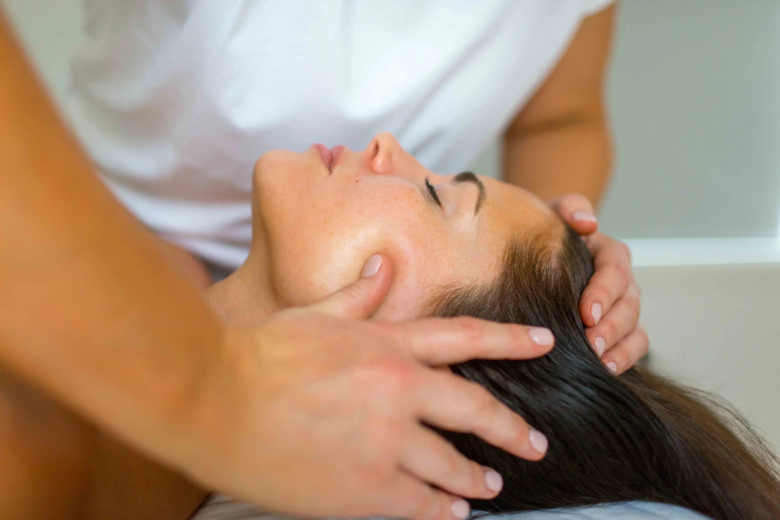 A person receiving a head massage with hands on their scalp and face, lying down with eyes closed.