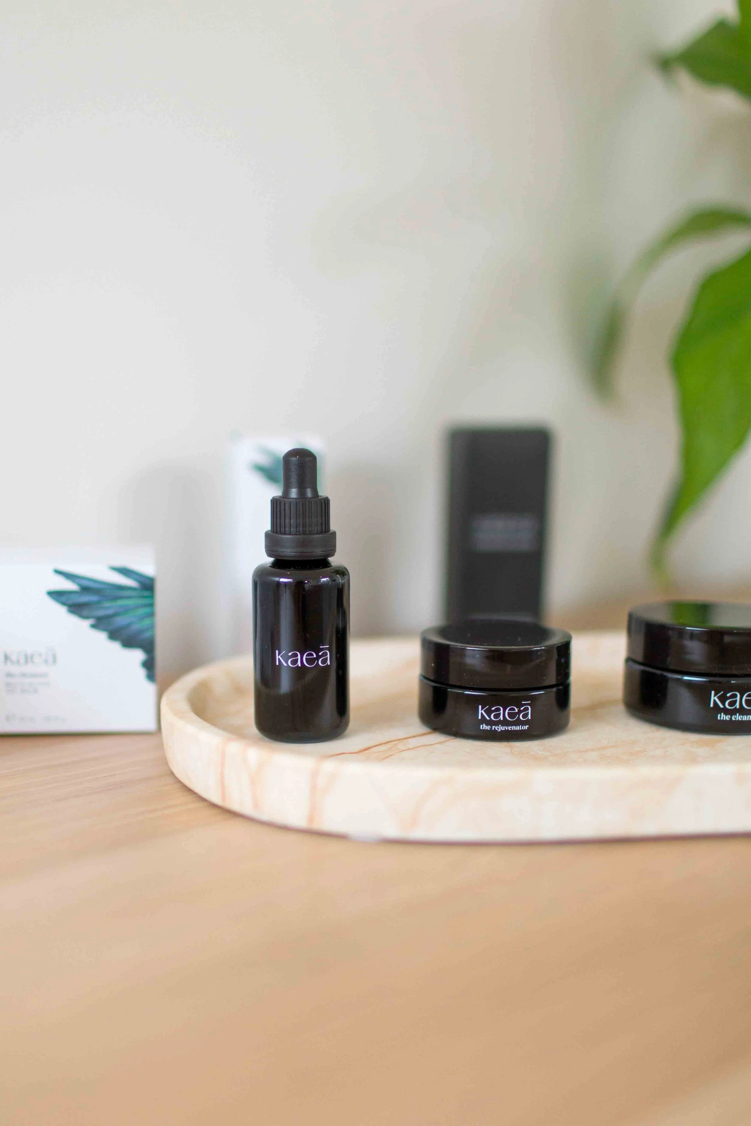 Skincare products on a wooden round tray, including a black dropper bottle labeled 'kaea', and two black jars labeled 'kaea the rejuvenator' and 'kaea the clean'. A white box with a green plant in the background.