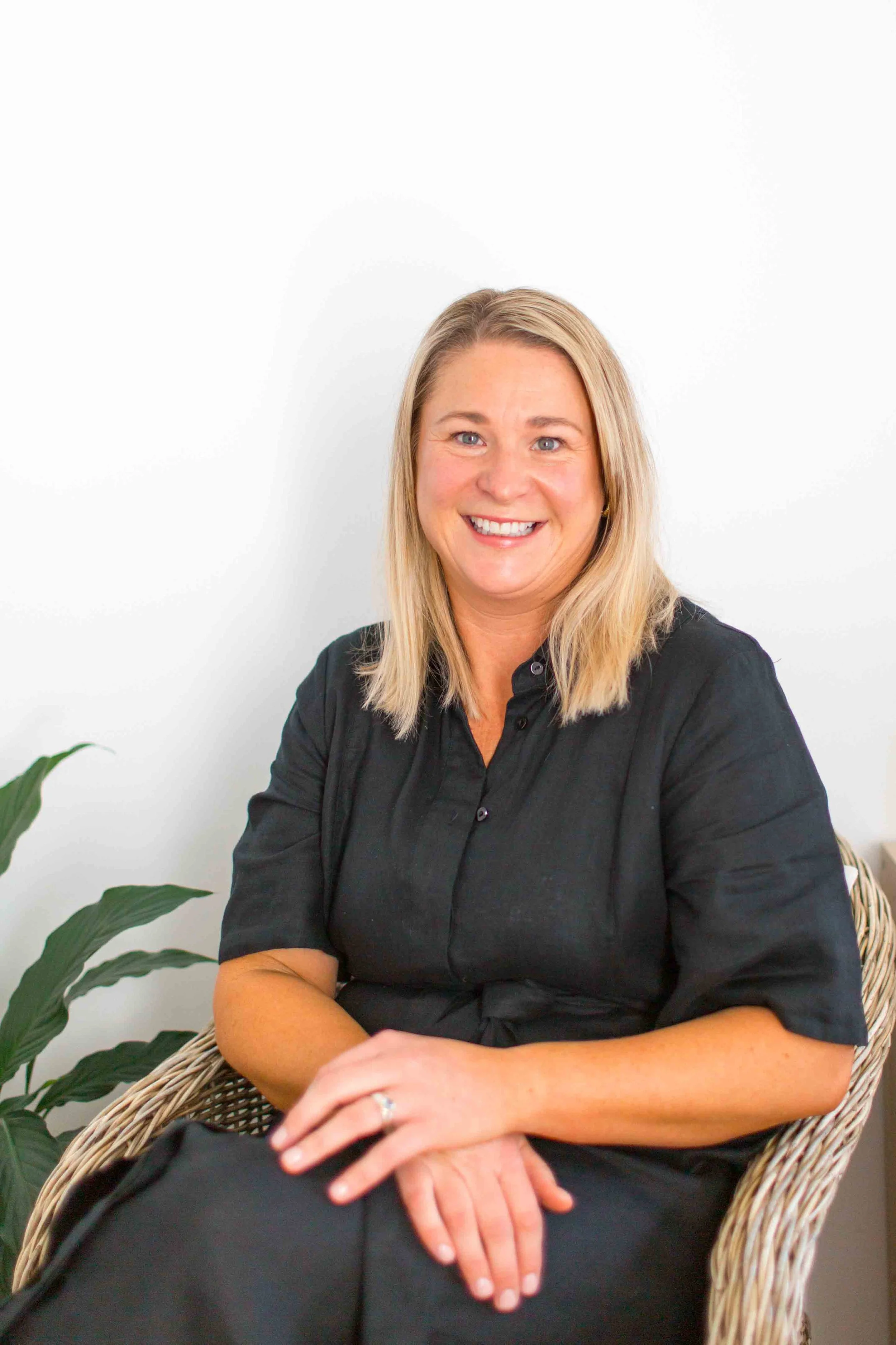 A woman with long blonde hair, wearing a black shirt, smiling while sitting on a wicker chair next to a green plant against a white wall.