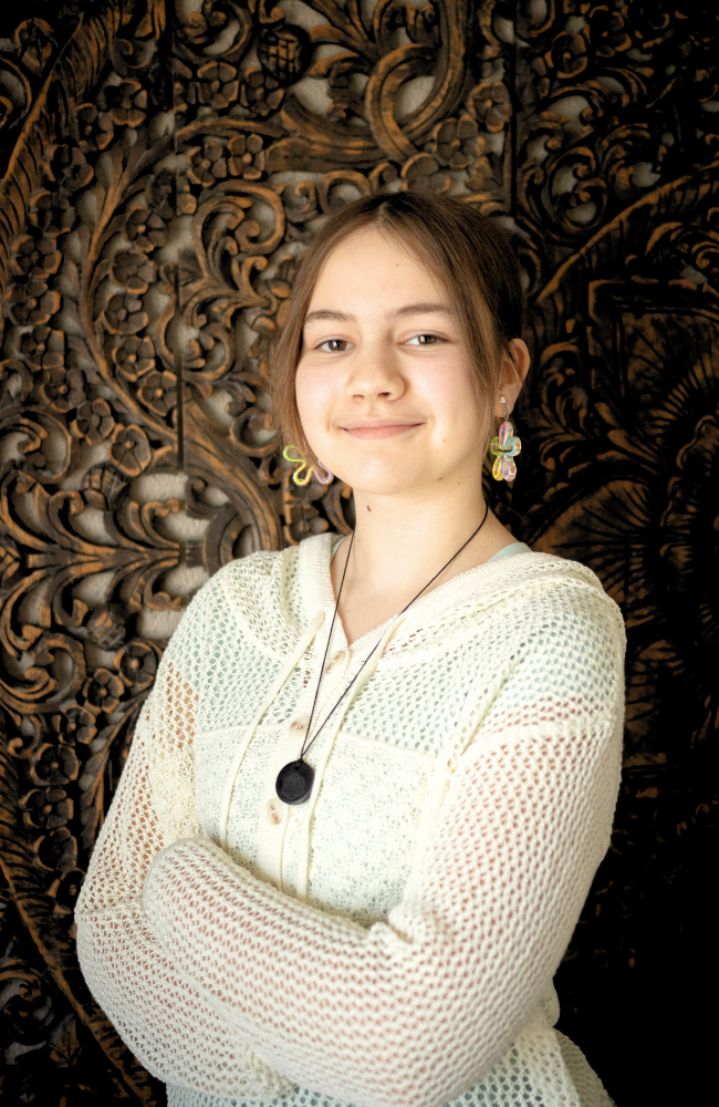 Young woman with earrings and a necklace, standing with arms crossed in front of an ornate carved wooden background.