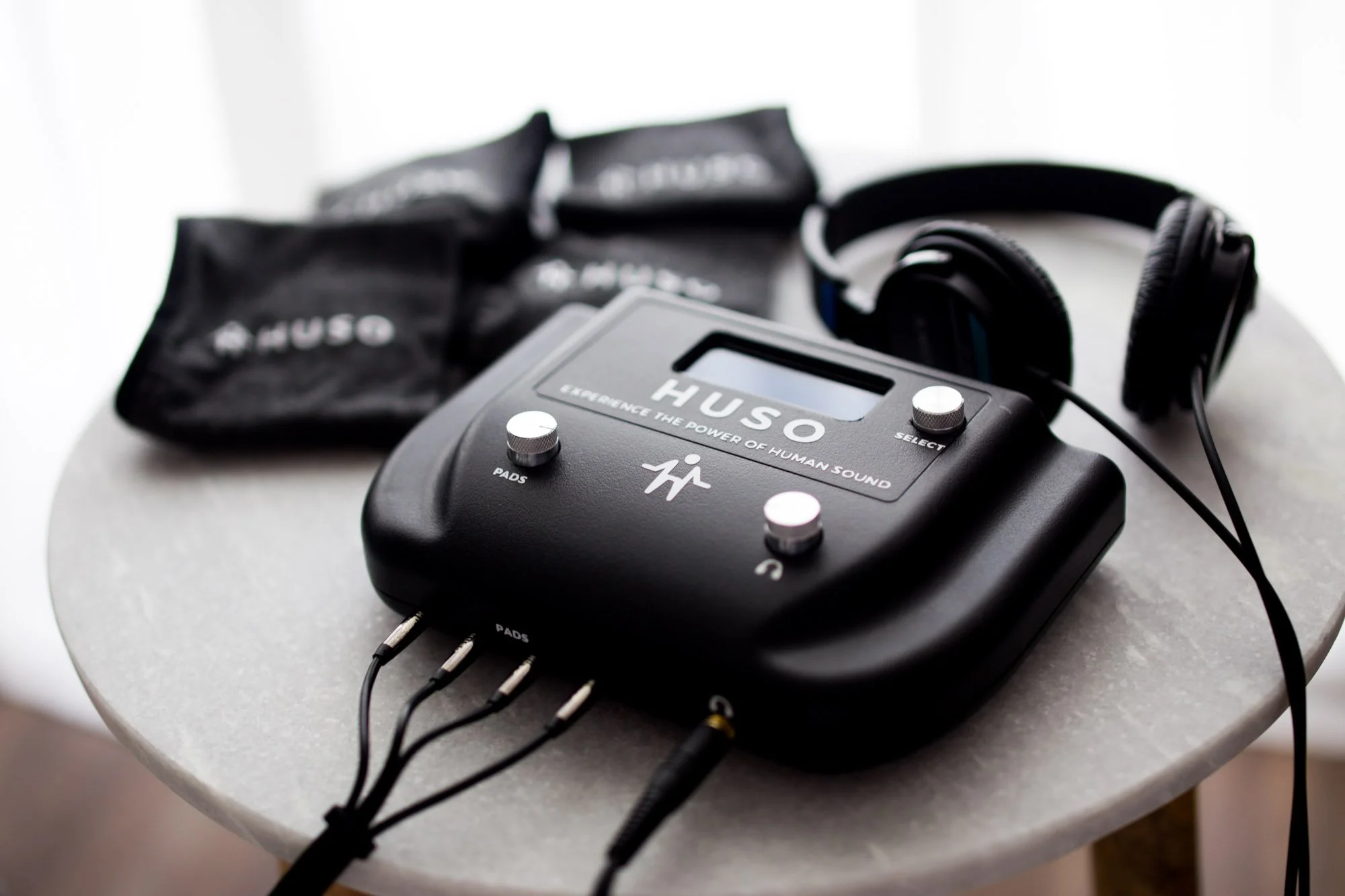 HUSO biofeedback device with wires connected to four pads on a round table, with black headphones and black bag in the background.