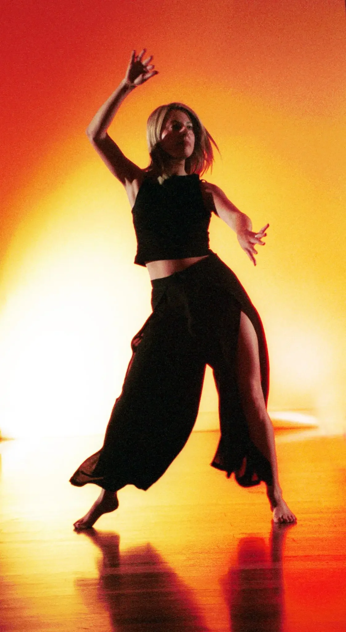 A woman dancing on stage with a yellow-orange background, wearing black clothing with a high slit, with one arm raised and the other extended to the side.