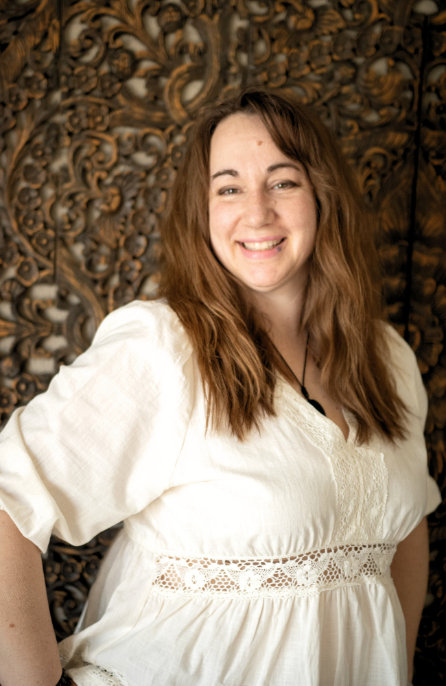 A woman with long, wavy brown hair smiling and wearing a white blouse with lace details, standing in front of an ornate wooden carved background.
