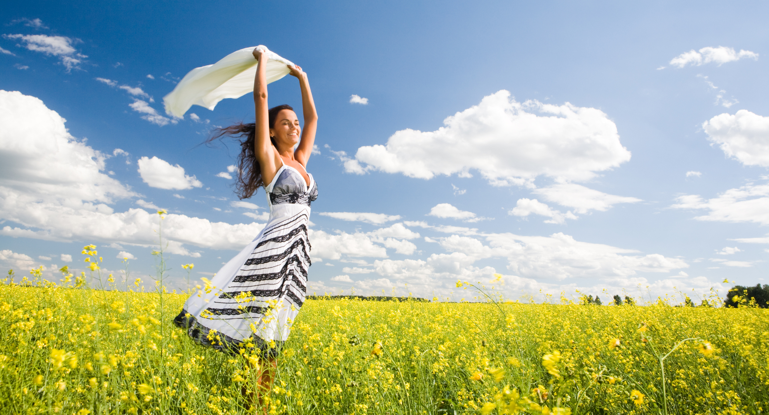 A woman in a white and black patterned dress standing in a yellow flower field, holding a white cloth above her head, smiling, with a blue sky and scattered clouds in the background.