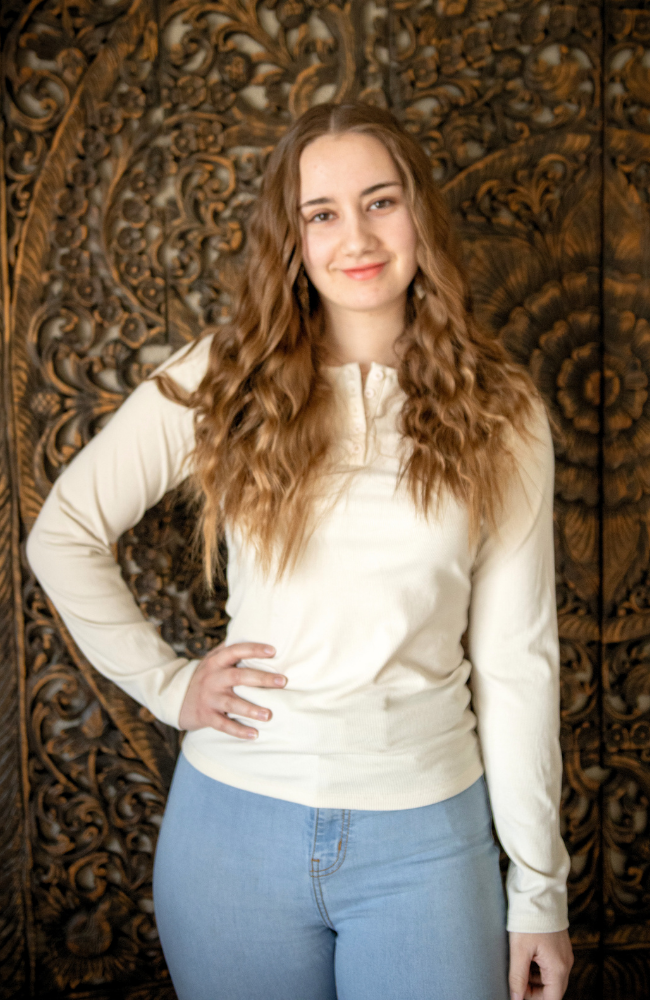 Young woman with long, wavy brown hair smiling and standing with one hand on her hip in front of a carved wooden background.