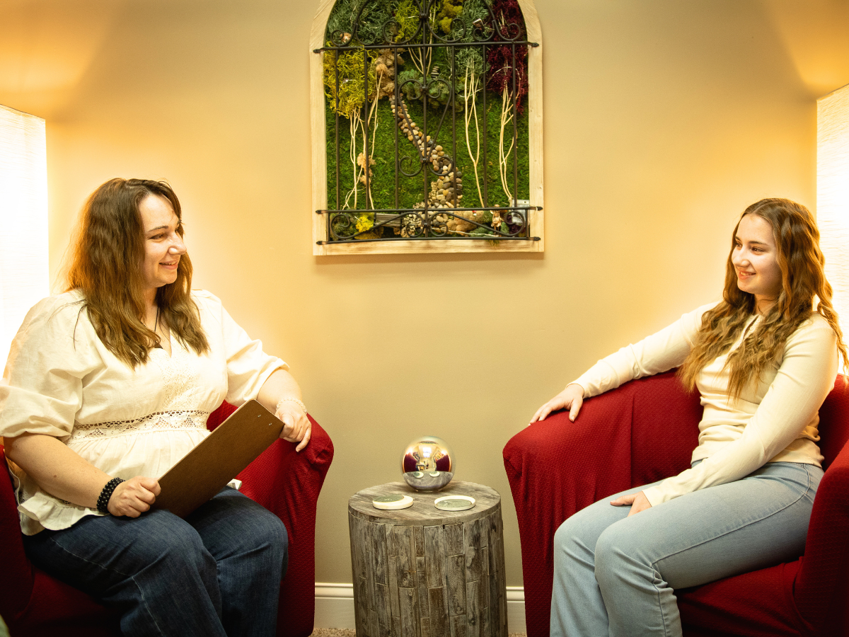 Two women sitting on red armchairs facing each other, smiling, in a cozy room with beige walls, a small round wooden table with decorative items, and a wall hanging with greenery and a heart-shaped design in the background.