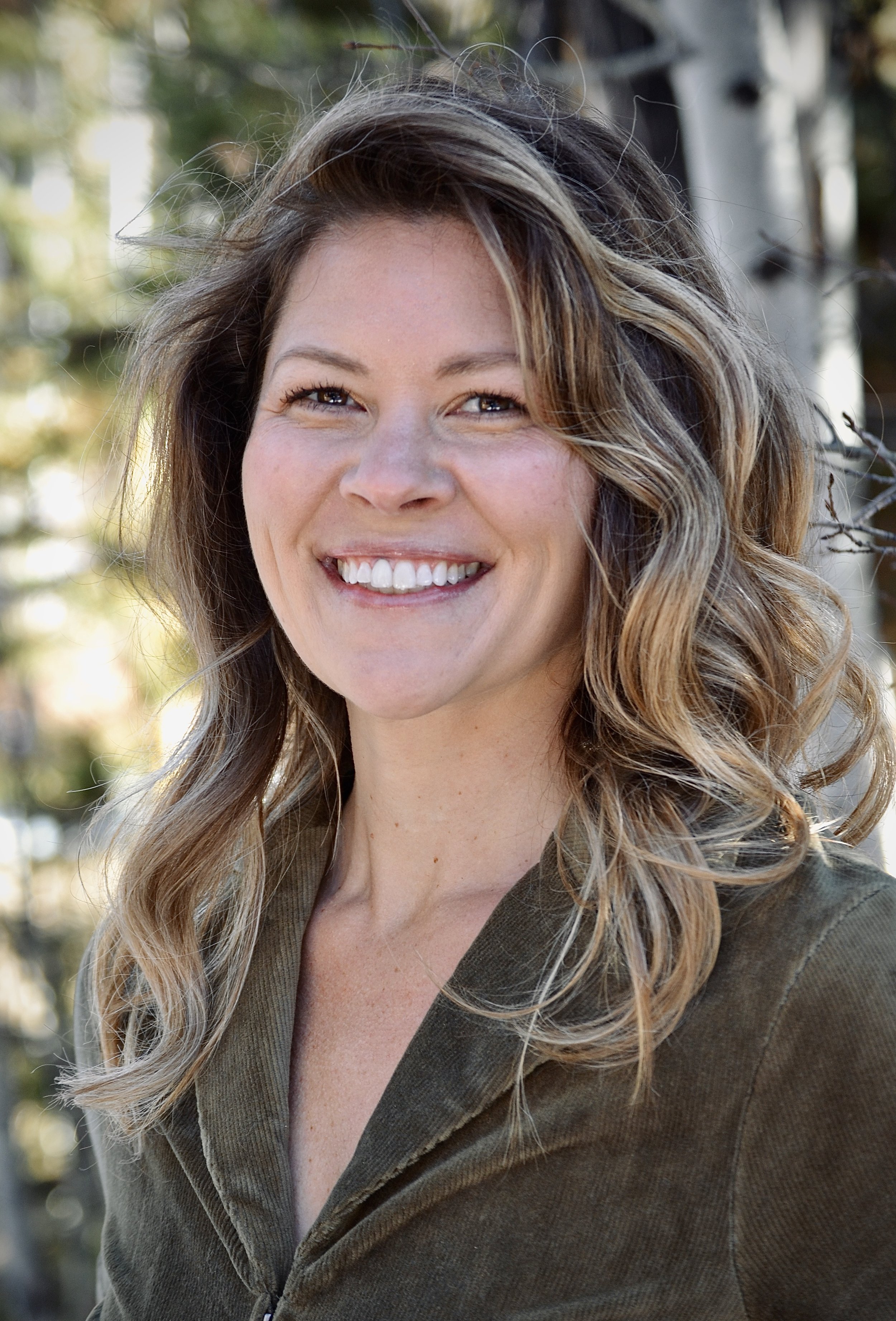 Close-up of a smiling woman with wavy, shoulder-length blonde hair outdoors with trees in the background.