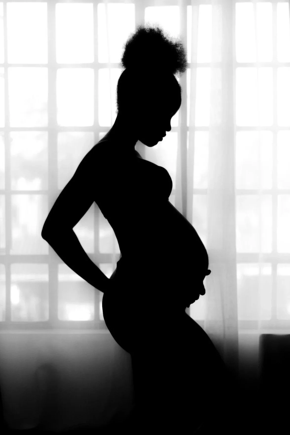 Silhouette of a pregnant woman standing in front of a window with sheer curtains.