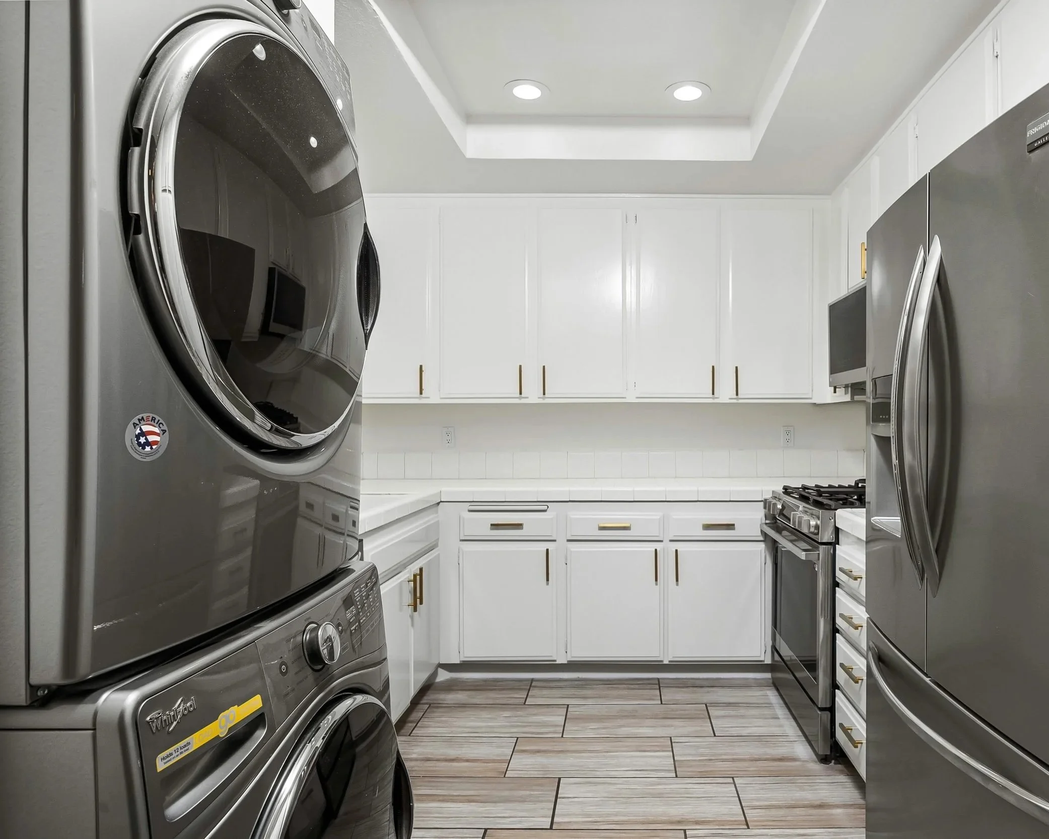 A modern laundry room with white cabinets, a front-loading washing machine and dryer, stainless steel refrigerator, microwave, oven, and tiled flooring.