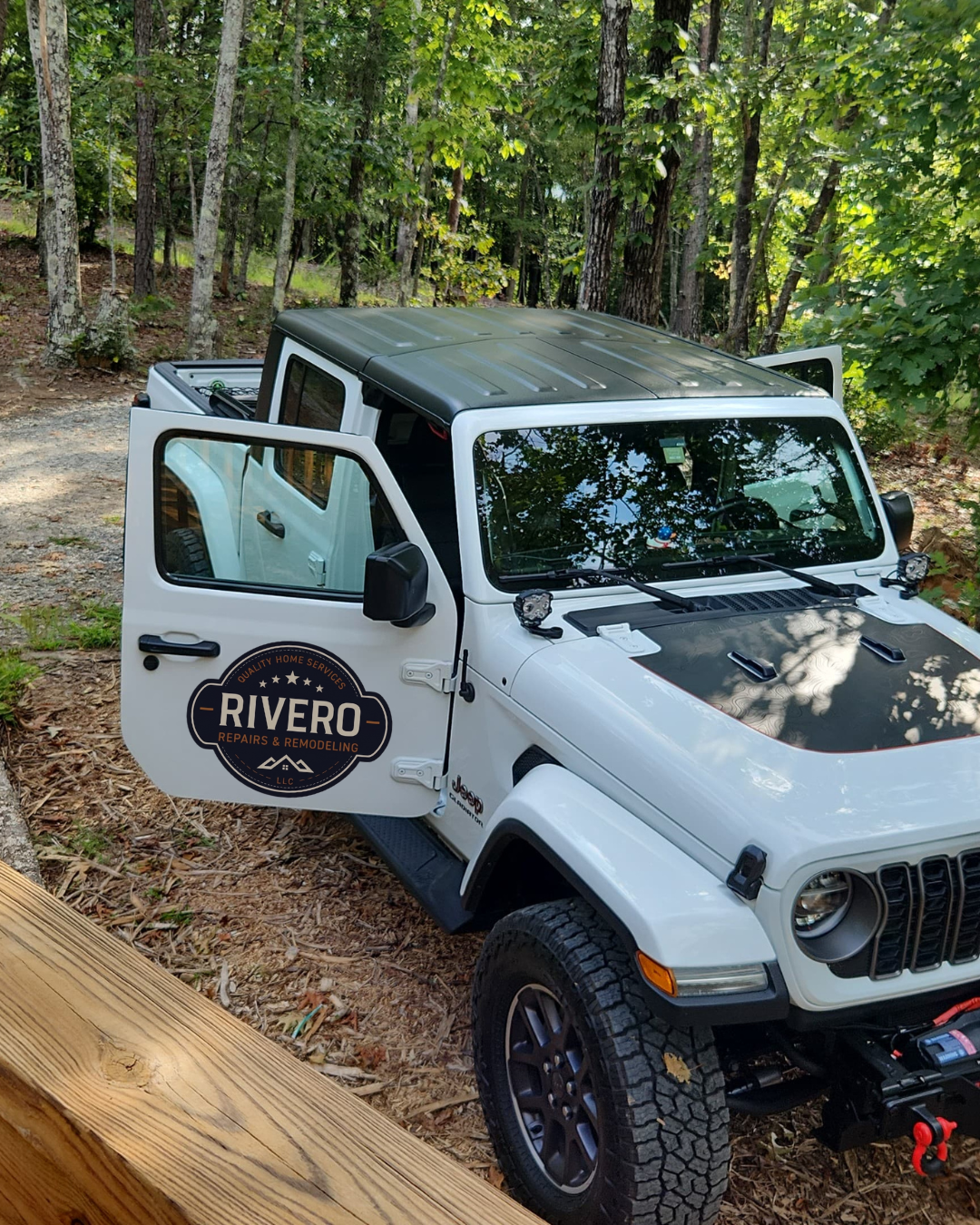 A white Jeep matched with a black roof parked on a dirt forest trail, with open driver’s side door and a logo for RIVERO REPAIRS & REMODELING on the door, surrounded by green trees. Jobsite Jeep. Cary. Home repairs and remodeling.