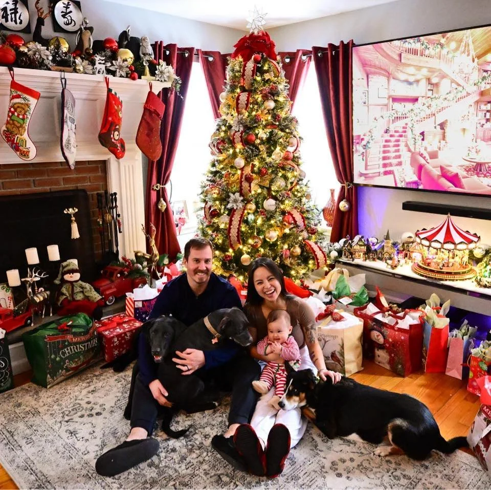 A family of four with two dogs sitting on the floor in front of a decorated Christmas tree in a living room, with wrapped gifts around them, stockings hanging on the fireplace, and Christmas decorations on the mantel.