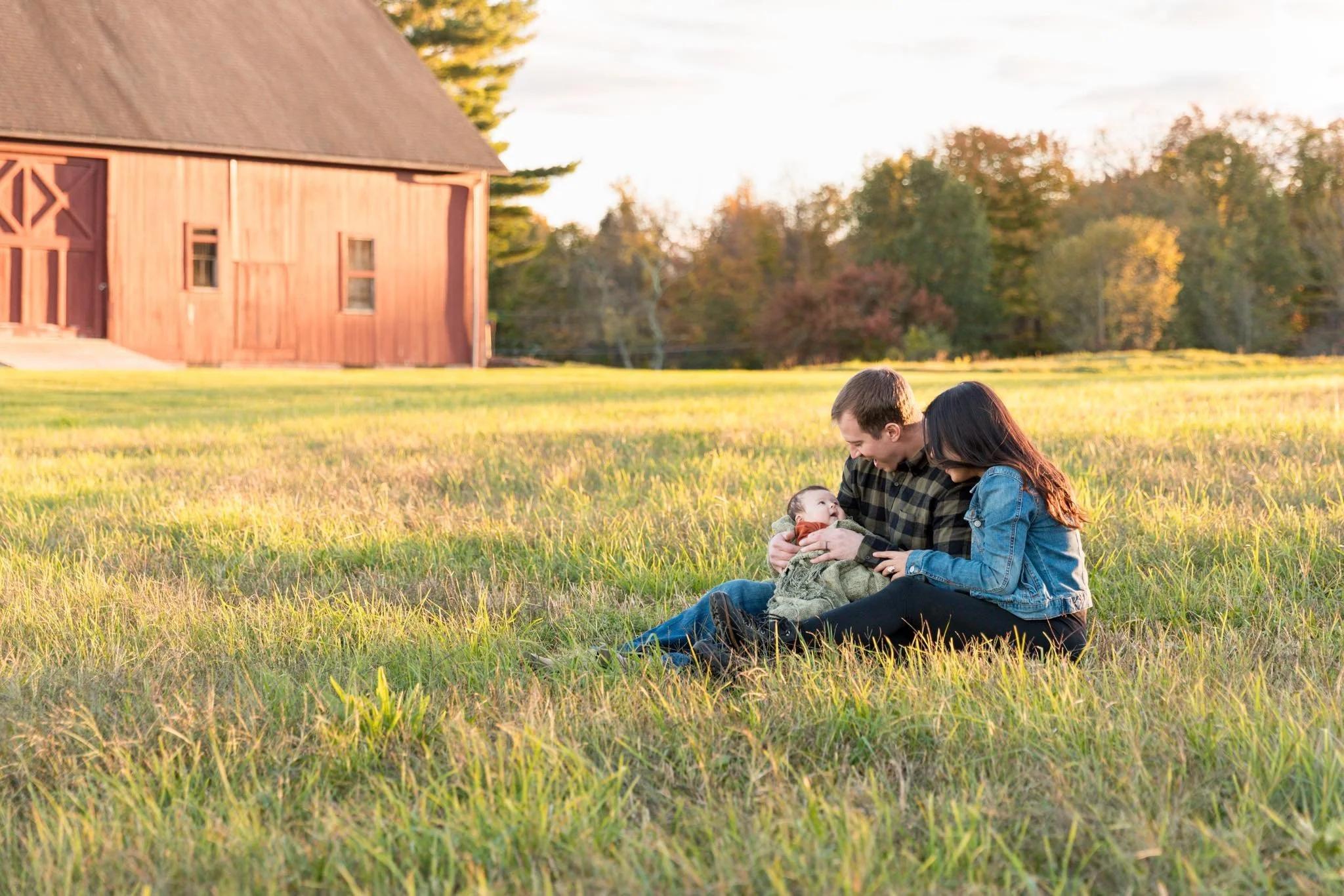 A family of three sitting on the grass in front of a red barn during sunset, sharing a moment with a baby.