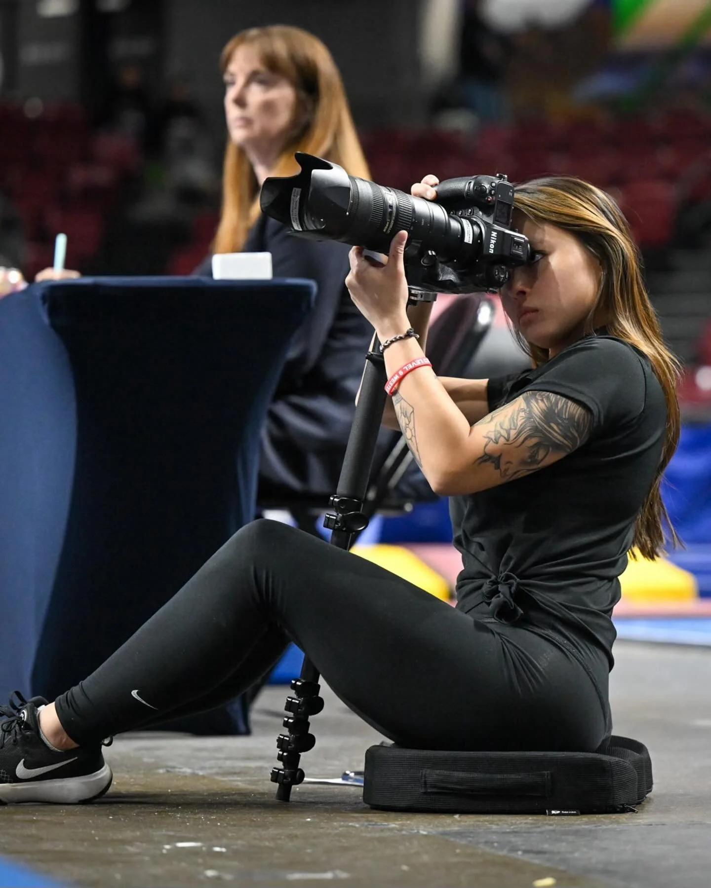 A woman with tattoos sitting on the floor, operating a professional camera mounted on a tripod, appear to be photographing or filming an event.