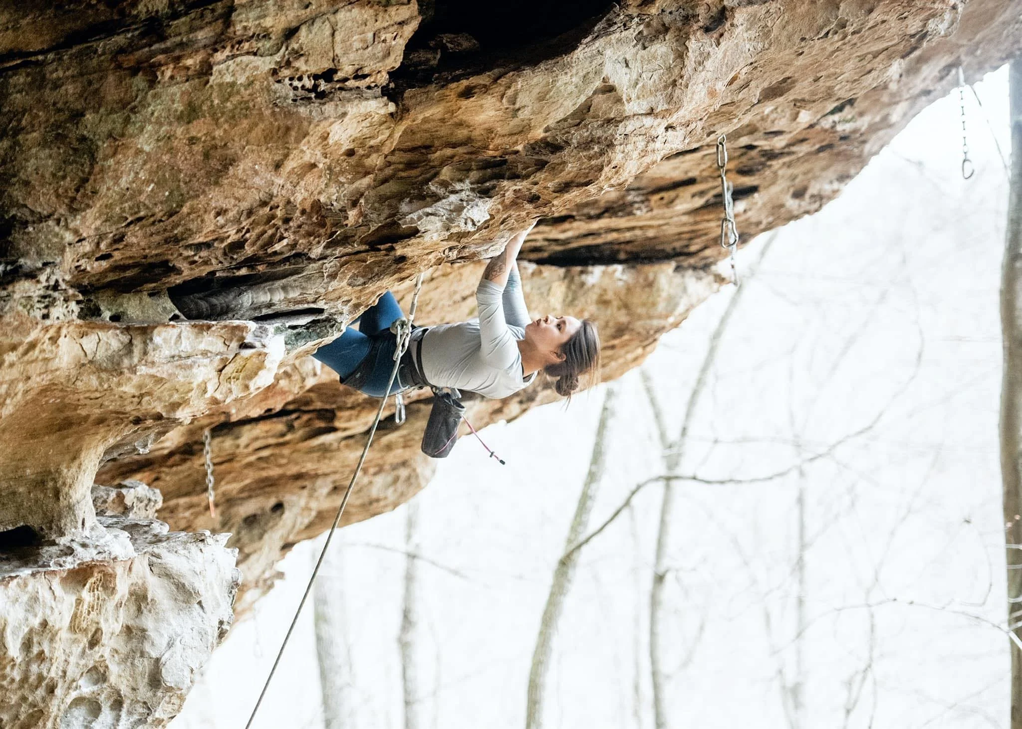 A woman rock climbing outdoors on a steep overhanging cliff, wearing a light-colored long-sleeve shirt, their hair in a bun, with climbing gear attached. The background shows trees and a cloudy sky.