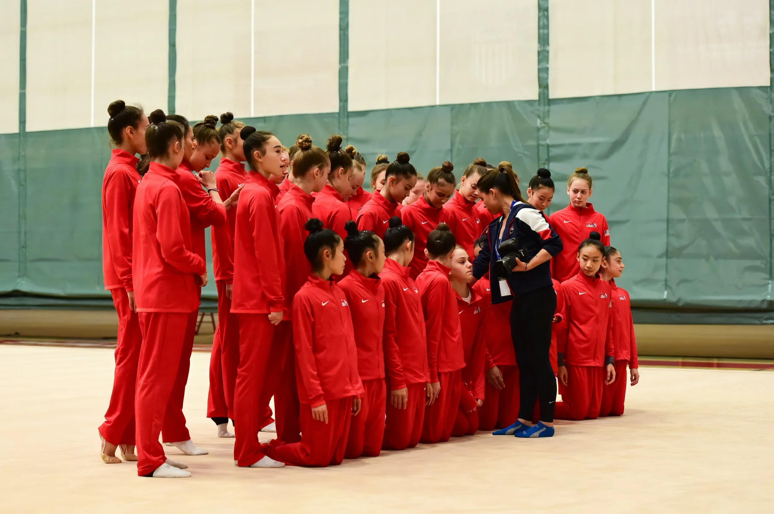 A group of female gymnasts in matching red tracksuits kneeling and standing in formation while a coach appears to be giving instructions, in an indoor gym.