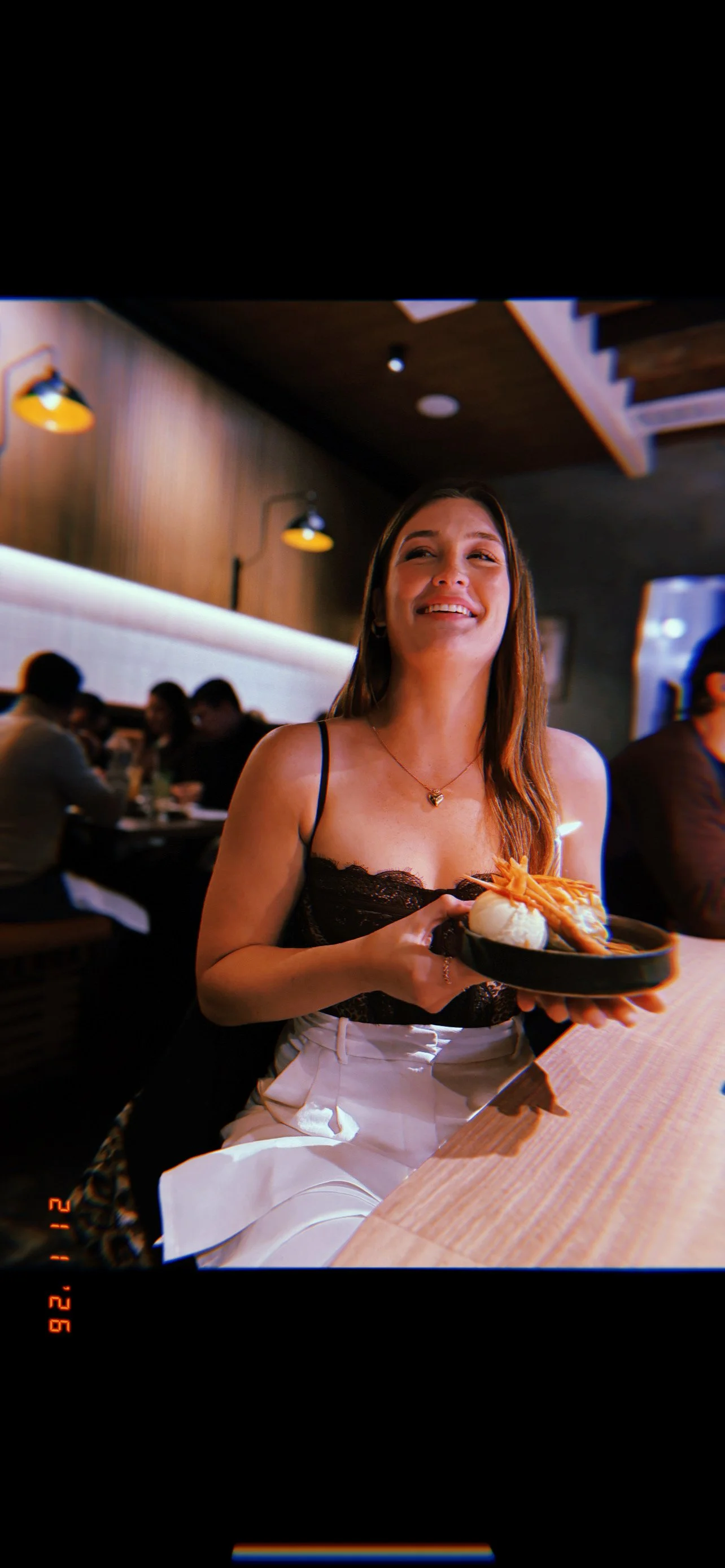A woman in a black lace top and white pants sitting at a restaurant table holding a plate of food, smiling. The restaurant has warm lighting, wooden accents, and other diners in the background.
