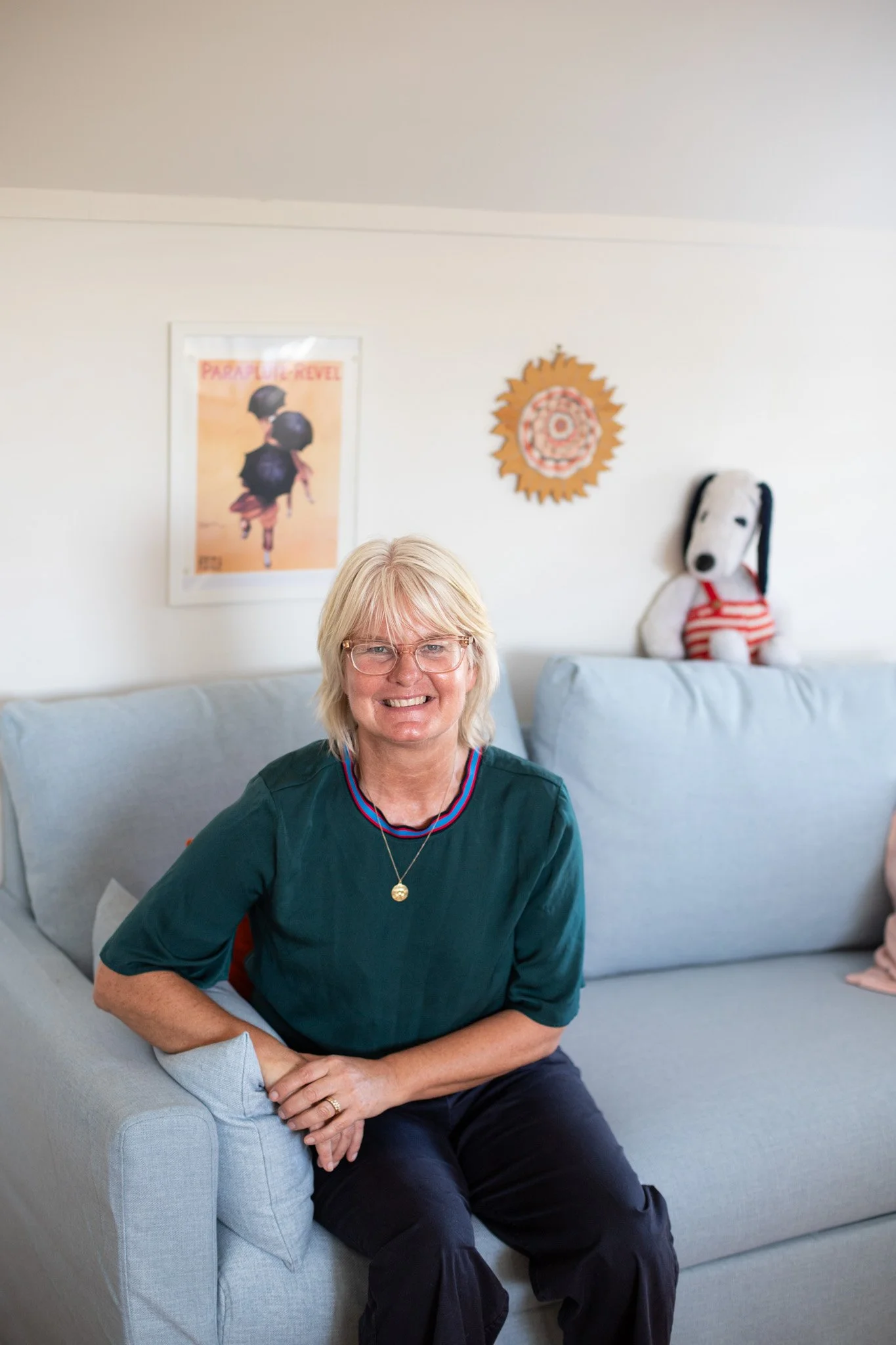 Tina sitting on a light blue couch, facing the camera and smiling. She is wearing a dark green tshirt, has blonde hair and wears glasses.