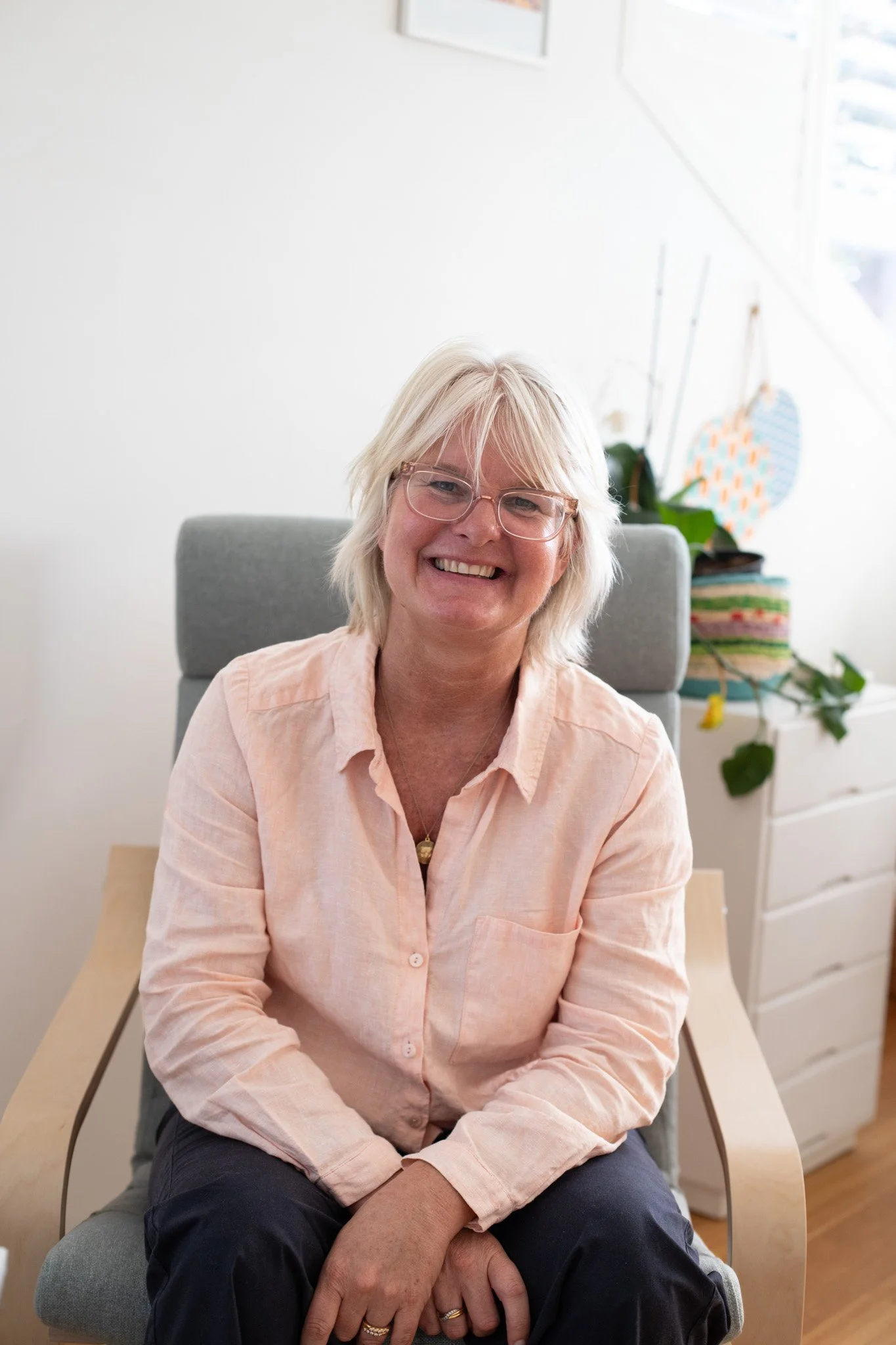 Tina is facing the camera and smiling while sitting in a grey arm chair. She is wearing a salmon coloured collared shirt and glasses. Her hair is blonde.
