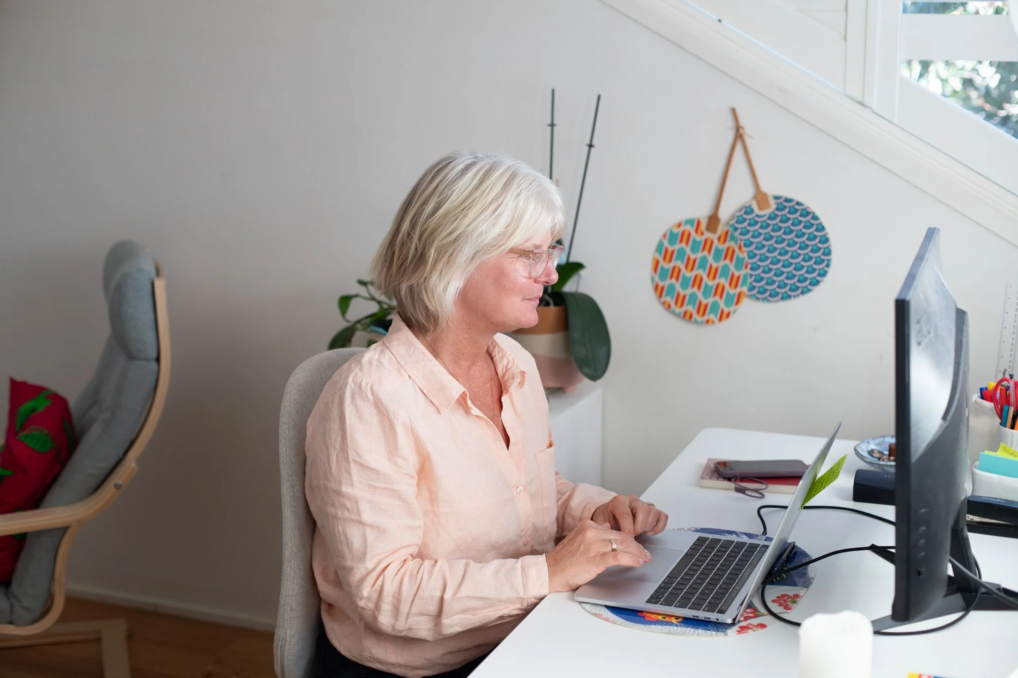 Tina is sitting at a desk on her laptop. She is wearing a salmon coloured collared shirt. She has blonde hair and glasses on.