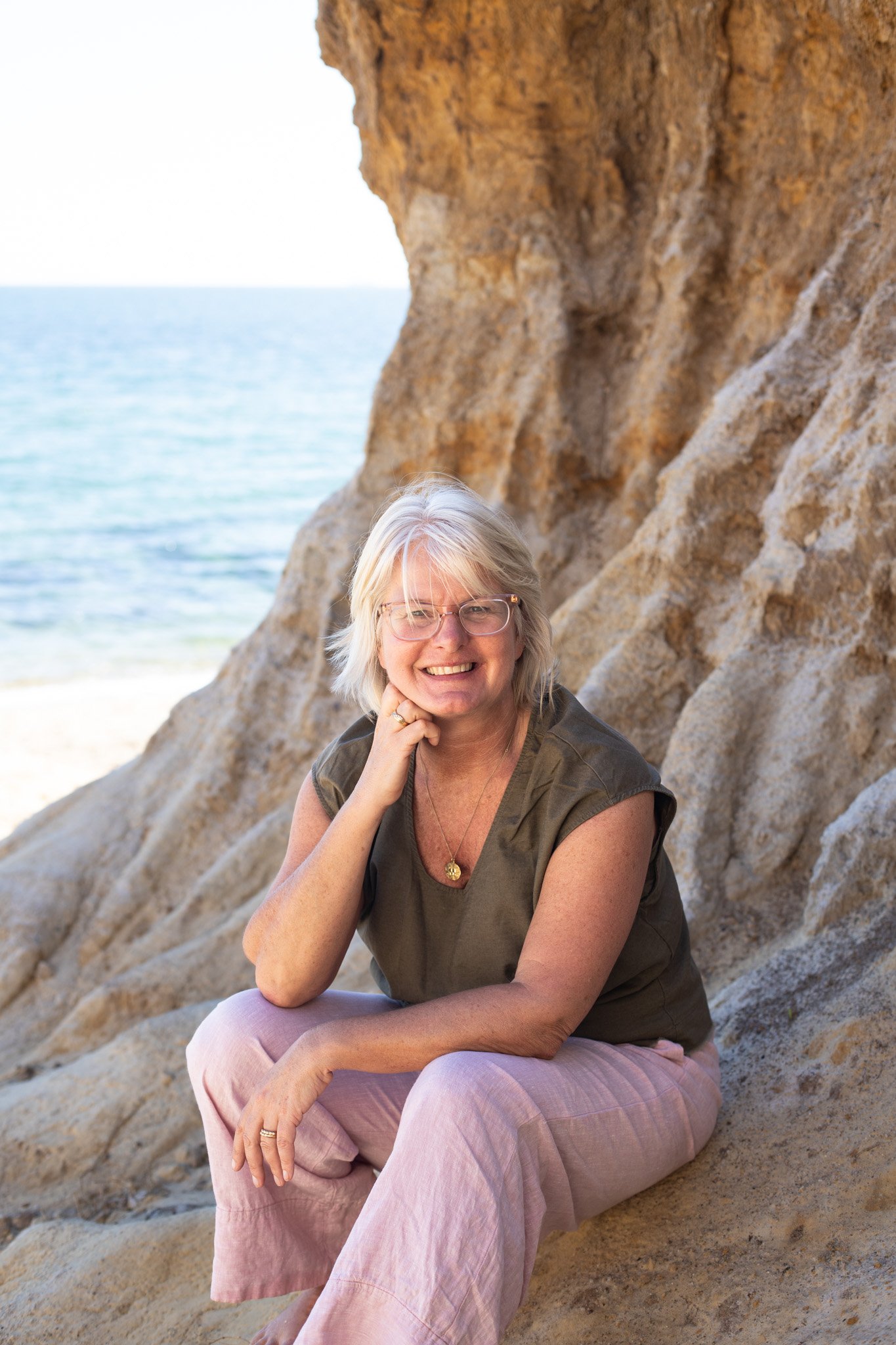 Tina is sitting on the side of a cliff on the beach, facing the camera and smiling. She has blonde hair and is wearing a kaki green tshirt, light pink linen pants and glasses.