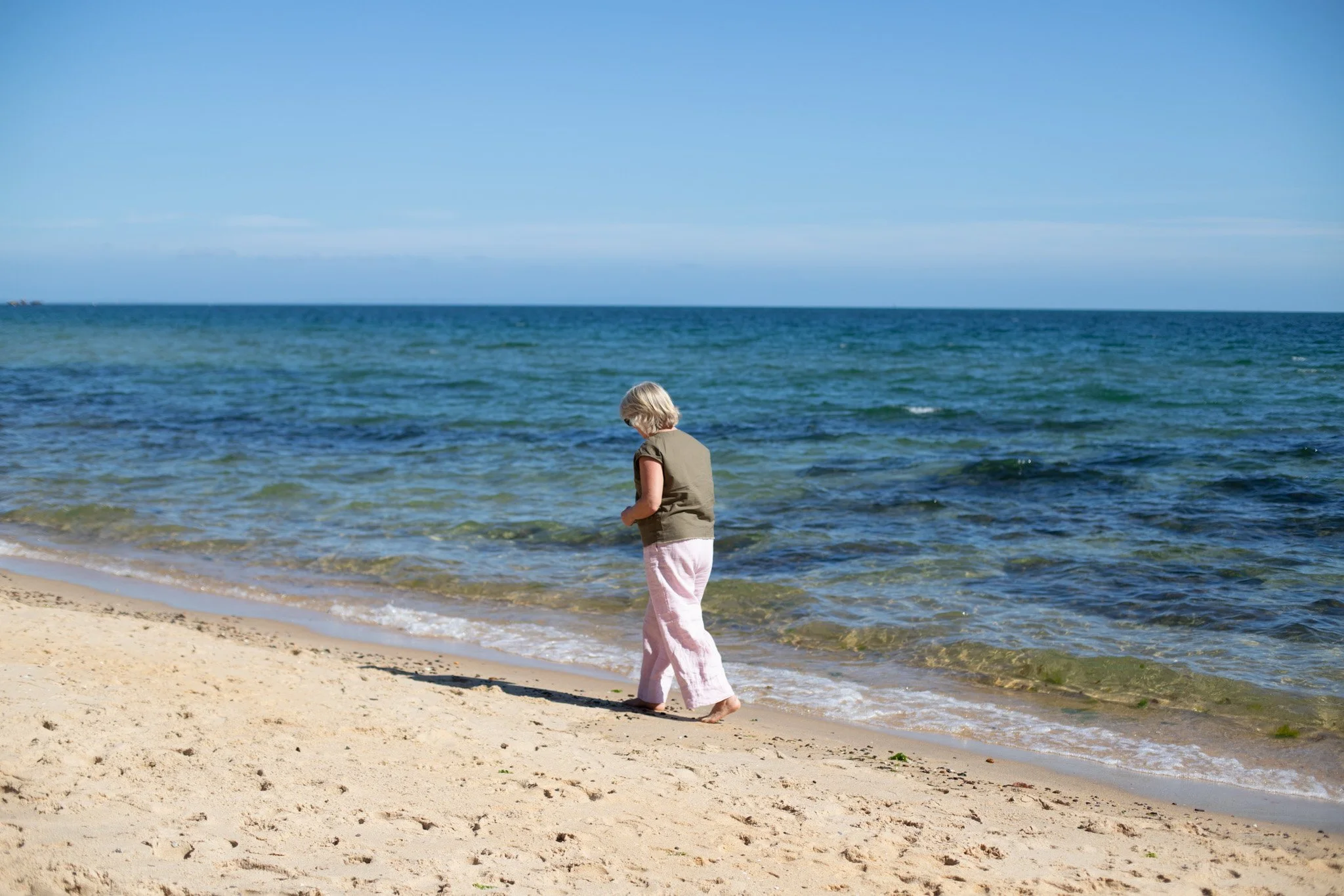 Tina is walking on the beach by the water. She is wearing a kaki green tshirt and white linen pants. She is facing away from the camera.