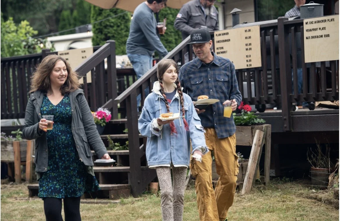 Family and friends enjoying outdoor food and drinks at a backyard gathering, with a woman holding a glass, a girl and man carrying plates of food.
