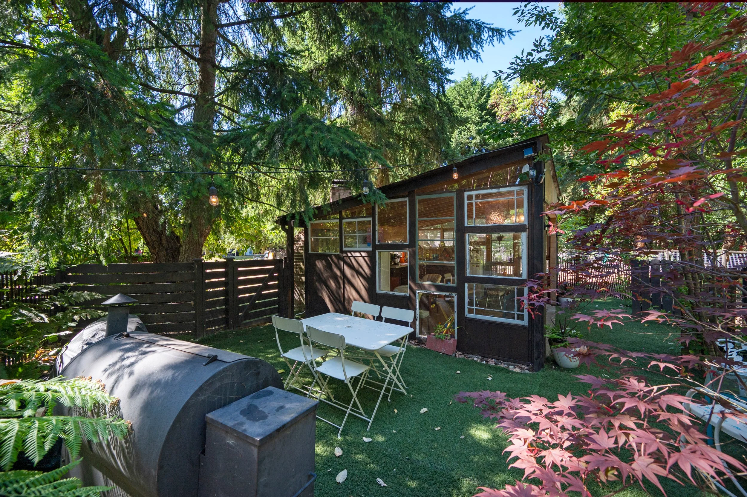 A backyard patio area with green trees and plants, a sunlit greenhouse or small structure with glass windows, outdoor white dining table and chairs, and string lights hanging above, surrounded by a wooden fence.