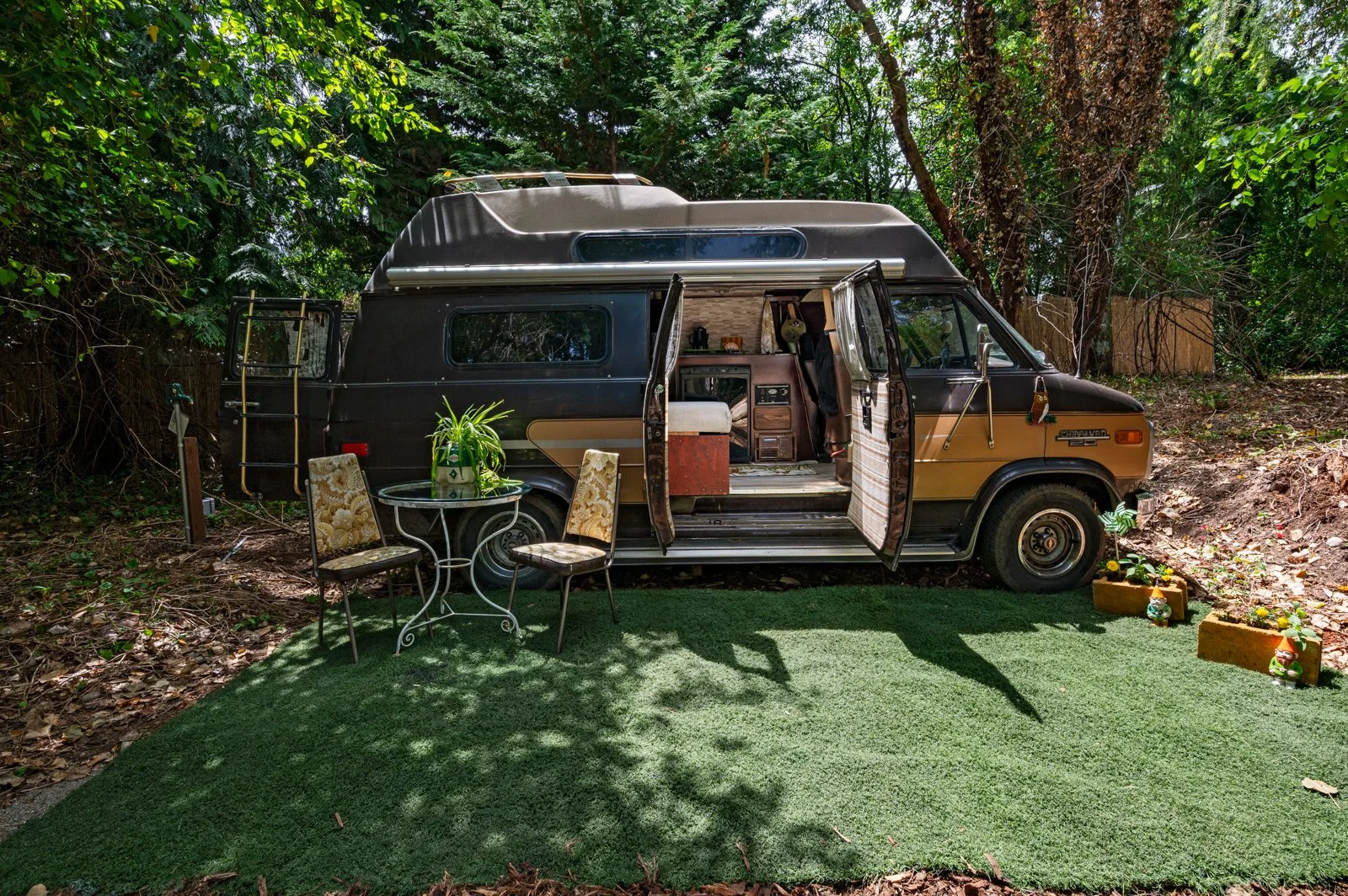 Vintage camper van parked in a wooded area with an outdoor patio setting, featuring a small table with chairs, potted plants, and decorative garden items on a small patch of artificial grass.