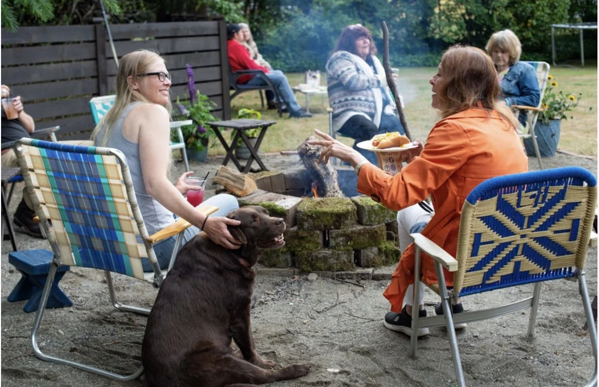 A group of women sitting around a backyard fire pit, socializing and enjoying food and drinks. One woman is petting a brown dog sitting on the ground.