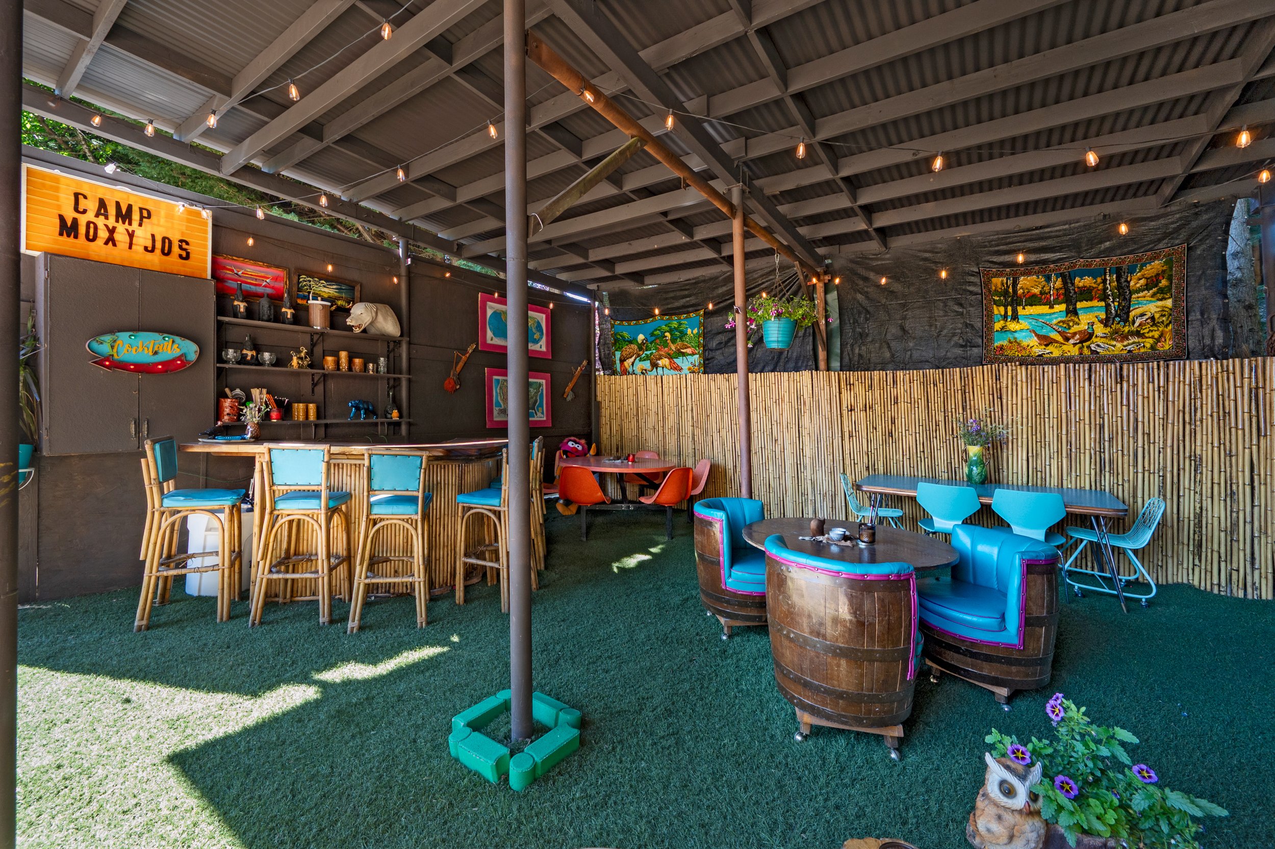 Outdoor seating area with colorful chairs, barstools at a wooden counter, hanging plants, wall art, and string lights at Camp Moxy Joes