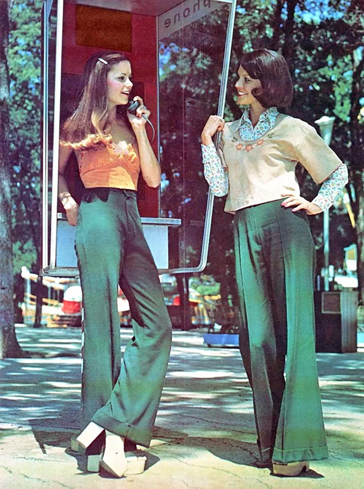 Two women with vintage hairstyles and clothing, standing outdoors near a red and glass structure. One woman is on a payphone, and the other is smiling and holding an umbrella. There are trees and parked cars in the background.