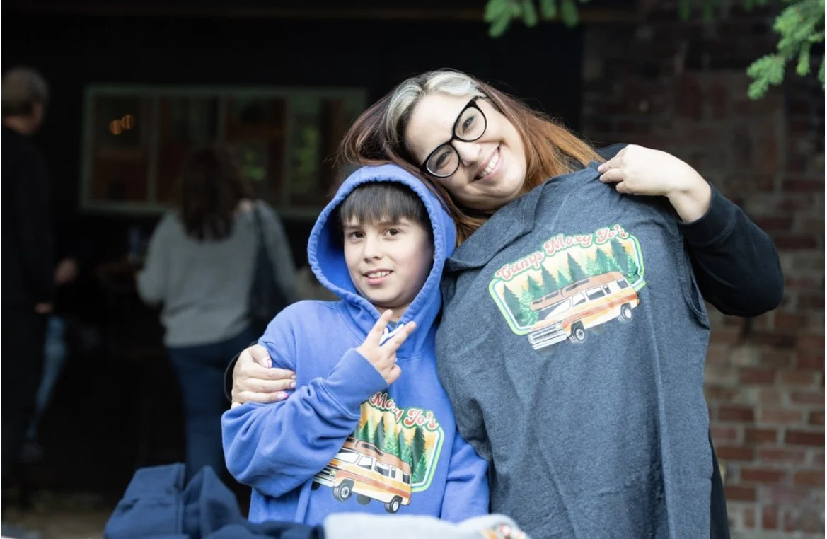 A woman with glasses and a young boy in a blue hoodie, both smiling, at a camp or outdoor gathering. The boy is making a peace sign. Both are wearing hoodies with a camper and forest graphic that says 'Camp Moxy Jo'.