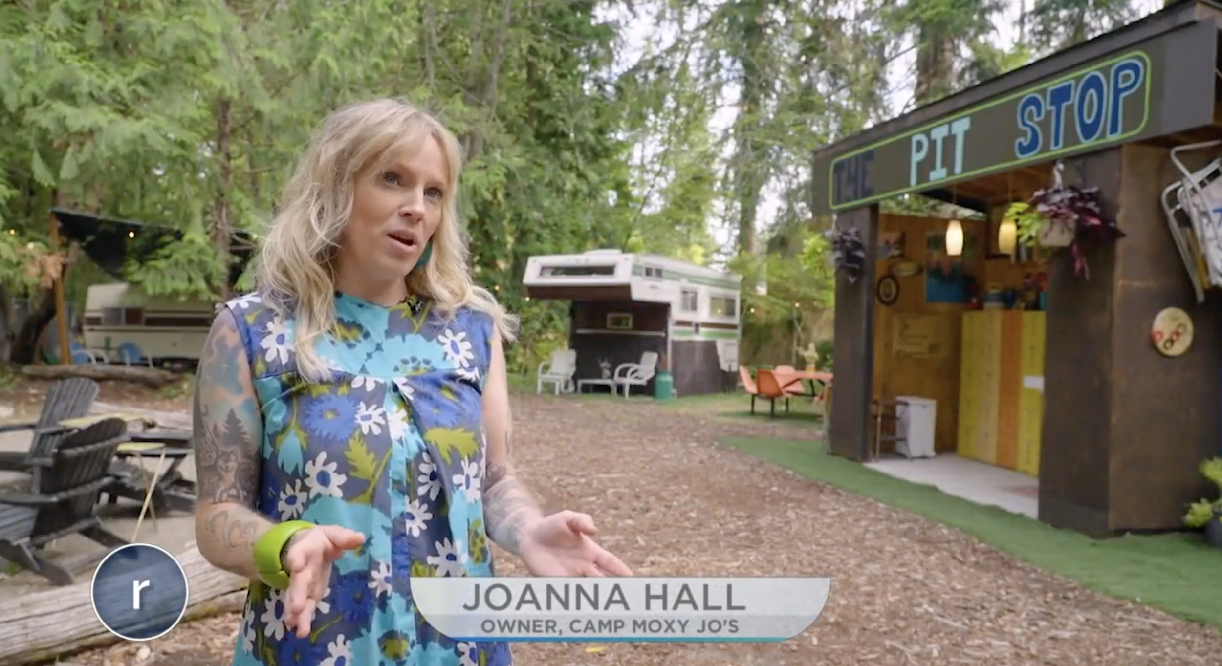 A woman standing outdoors at a campground, with a sign reading "The Pit Stop" on a small wooden building behind her. The woman has wavy blonde hair, tattoos, and is wearing a colorful floral dress. Trees and camping trailers are in the background.