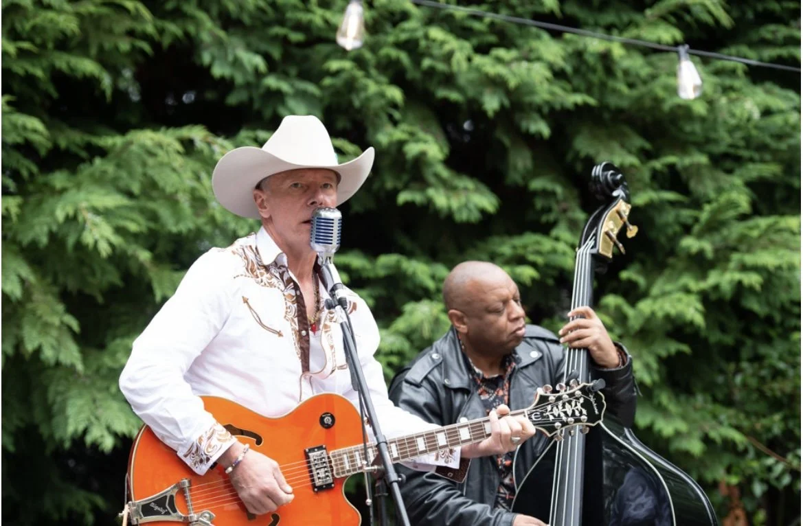 Two musicians performing outdoors, one playing guitar and singing into a microphone, the other playing upright bass, with green trees in the background.