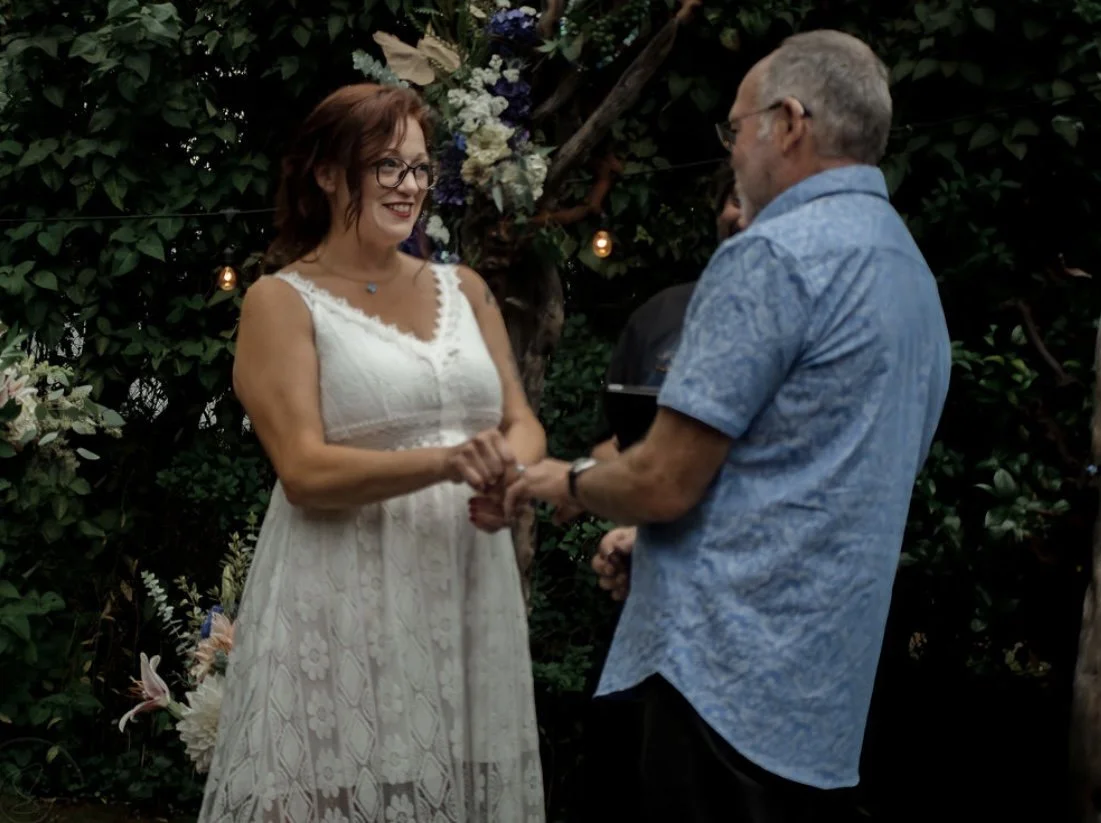 A woman in a white lace dress and glasses is holding hands with a man in a light blue shirt, during a wedding ceremony outdoors at night, surrounded by greenery and string lights.