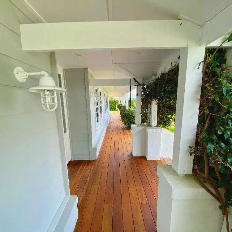 A wooden porch with white columns and a row of hedges along the side, featuring outdoor wall lights and a view of a garden in the distance.