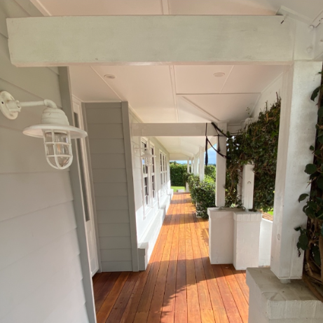 Covered porch with a wooden floor, white walls, and potted plants, overlooking a garden with green trees.