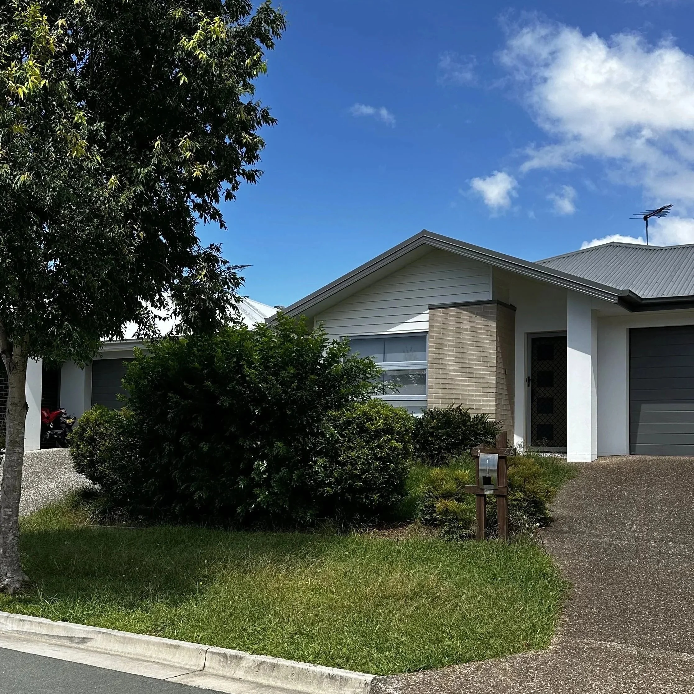 A modern single-story house with a driveway, surrounded by bushes and trees, under a blue sky with some clouds.