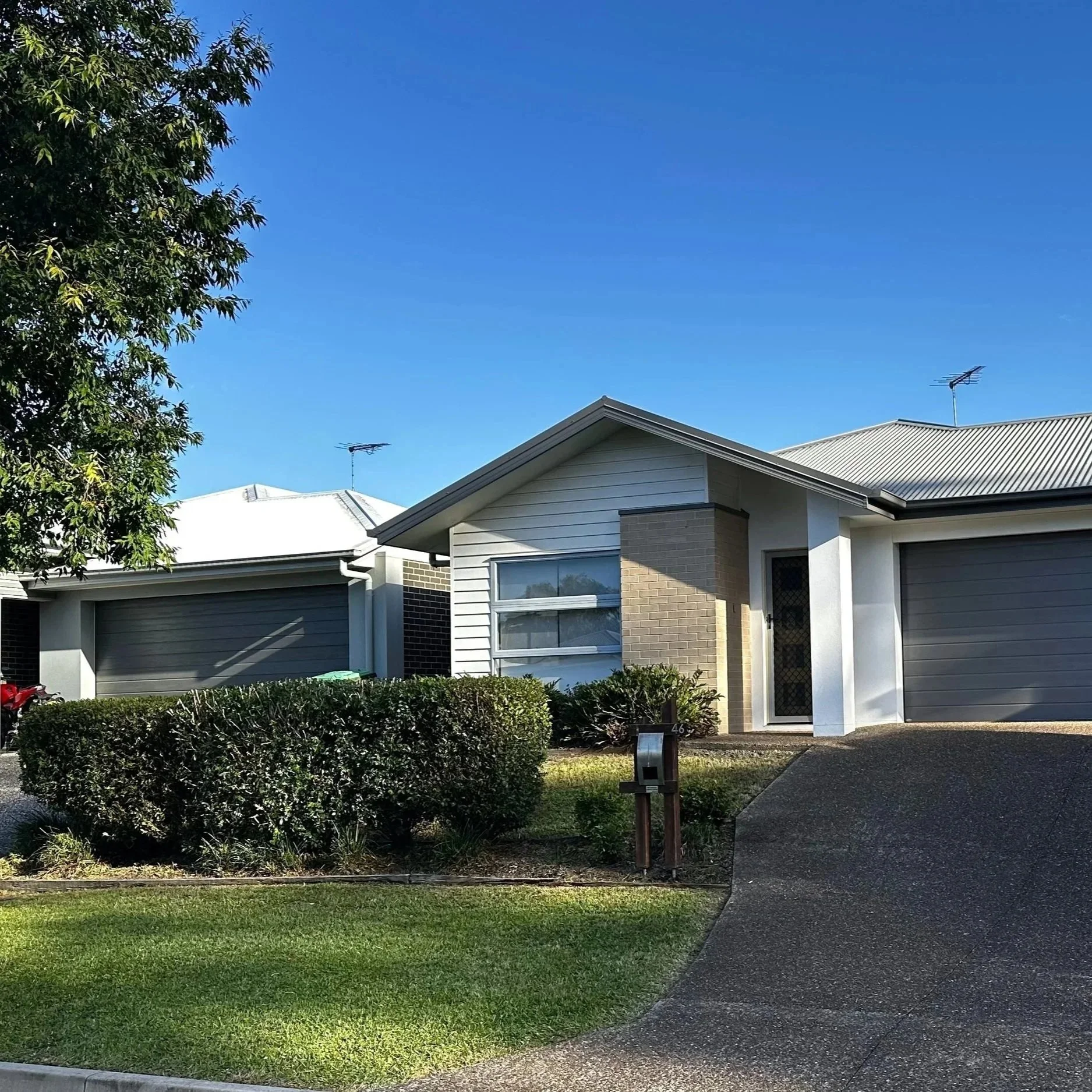 Front view of a suburban house with a driveway, green lawn, bushes, and a tree, under a clear blue sky.