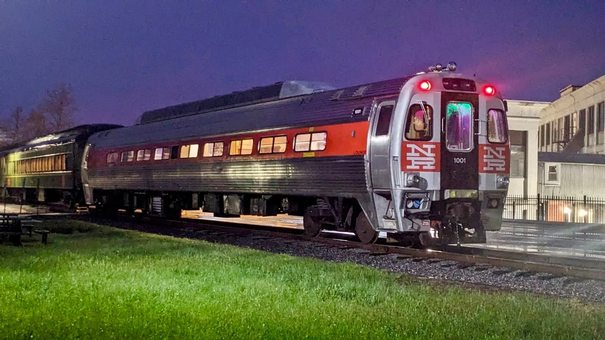 A vintage silver train car with red accents and the number 1001 on the front, parked on train tracks at dusk with a purple sky and a building in the background.