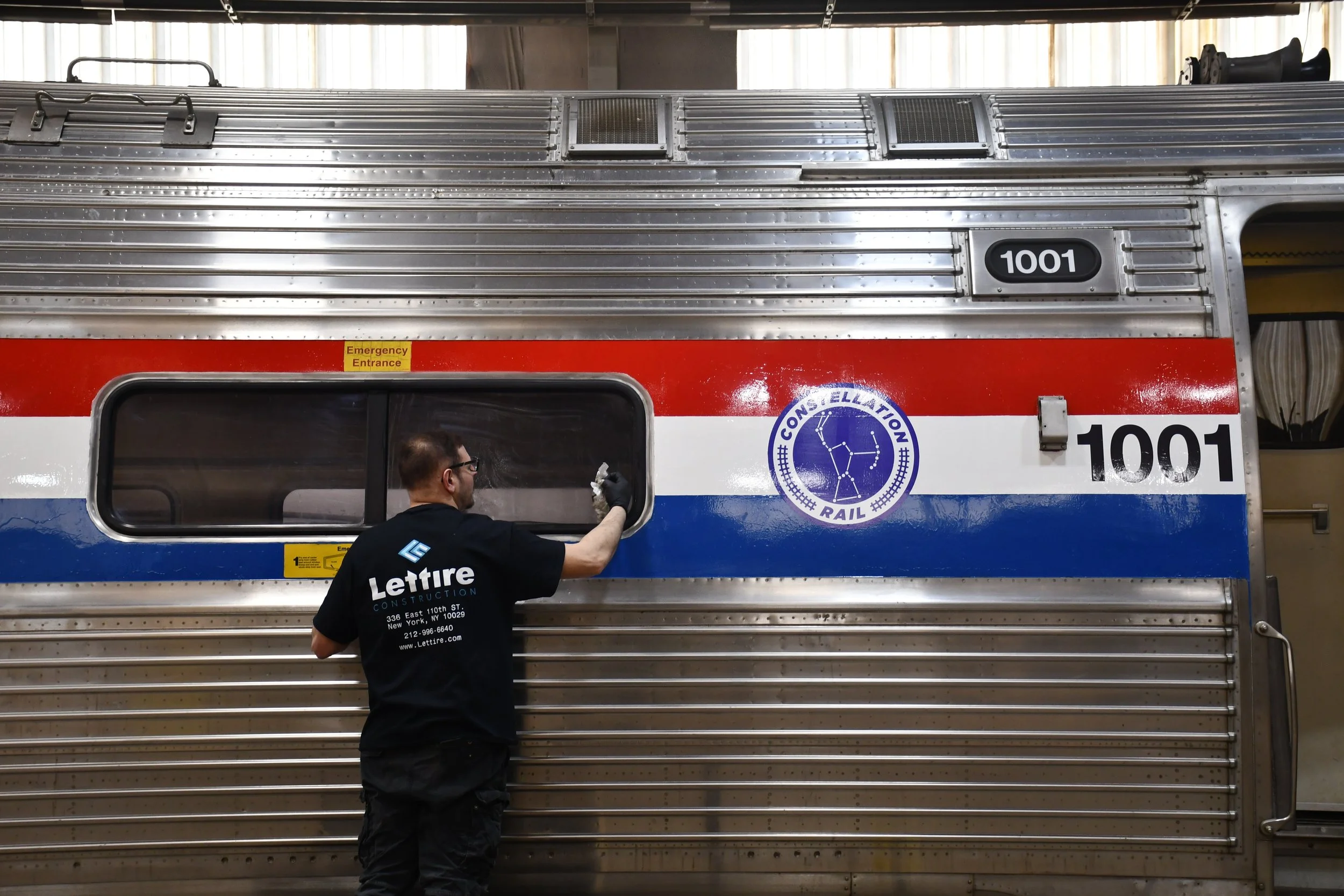 A worker cleaning the window of a silver, red, blue, and white train car with the number 1001 and a constellation rail logo, inside a maintenance facility.