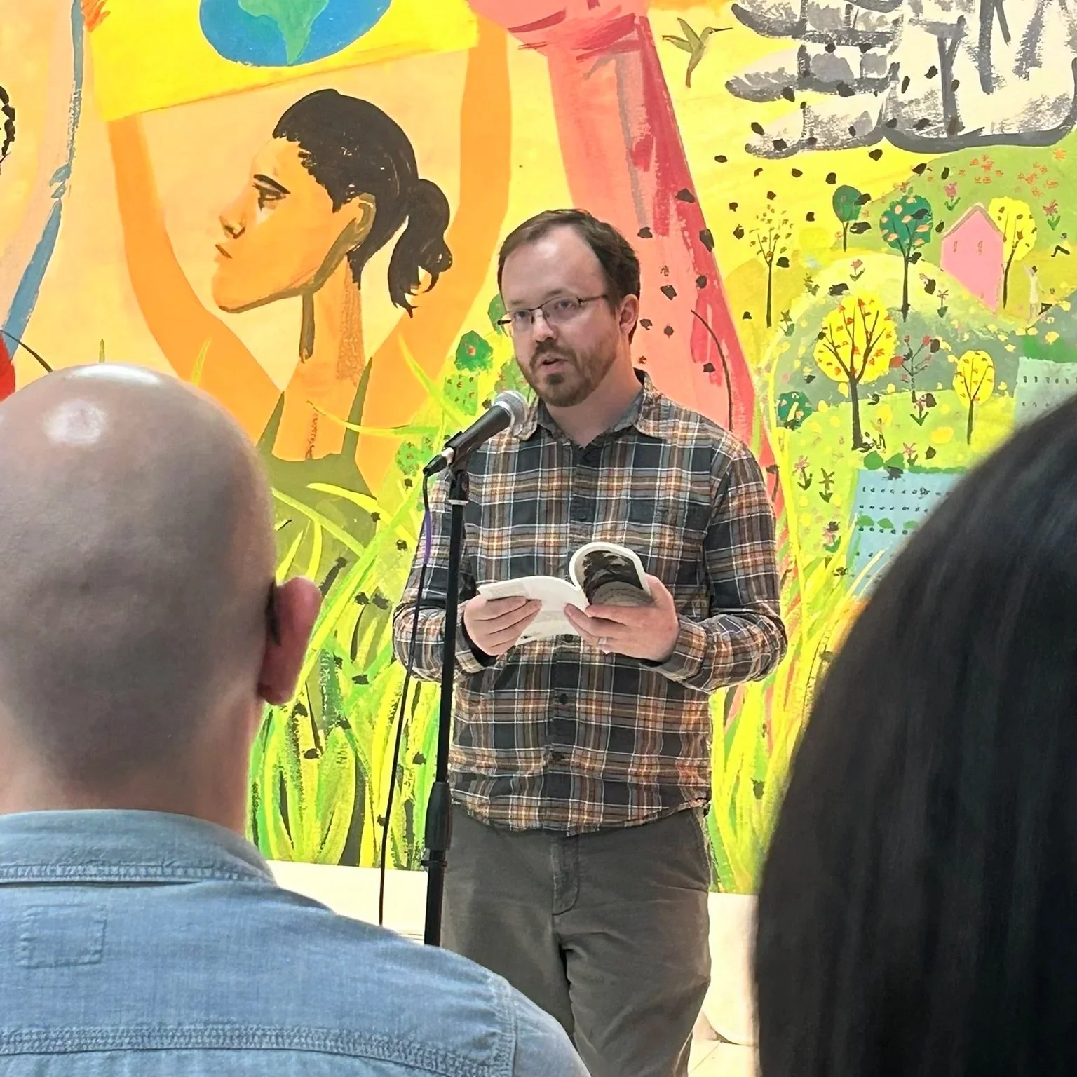 A man in his mid-30s, identified as John Linstrom, stands at a microphone holding an open poetry book from which he appears to be reading. A colorful mural spans the wall in the background.