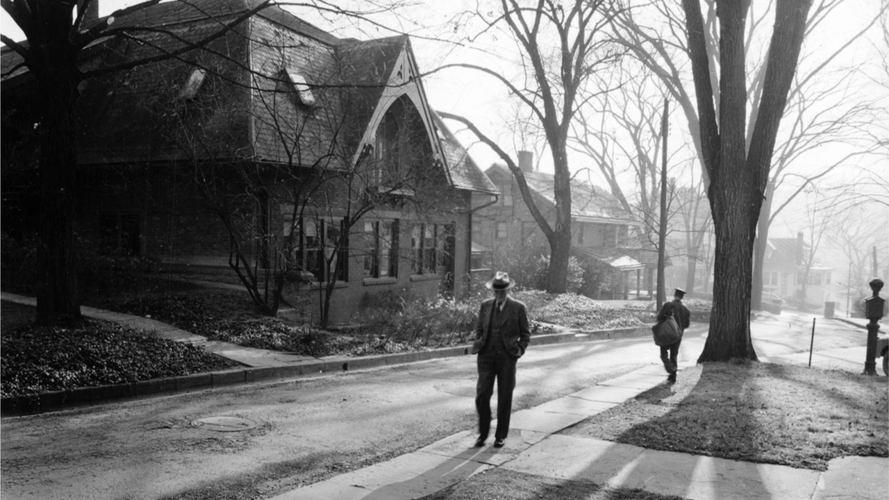 Detail of a late-life photograph of Liberty Hyde Bailey walking in front of his now-historic property on Sage Place, Ithaca, NY. An evening shadow stretches in front of Bailey along the sidewalk.
