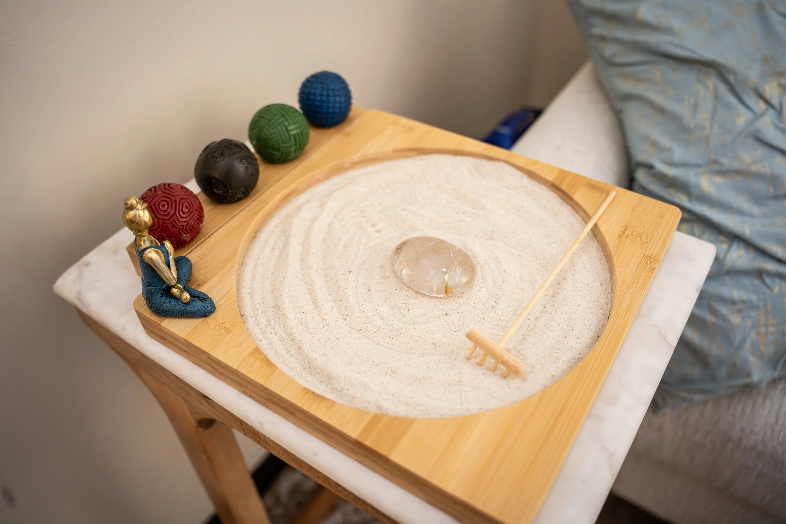 A wooden tray with sand and a large, clear, rounded crystal in the center. There are five colorful textured spheres along one edge of the tray, and a small figure of a person sitting with their hands on their head near the sphere.