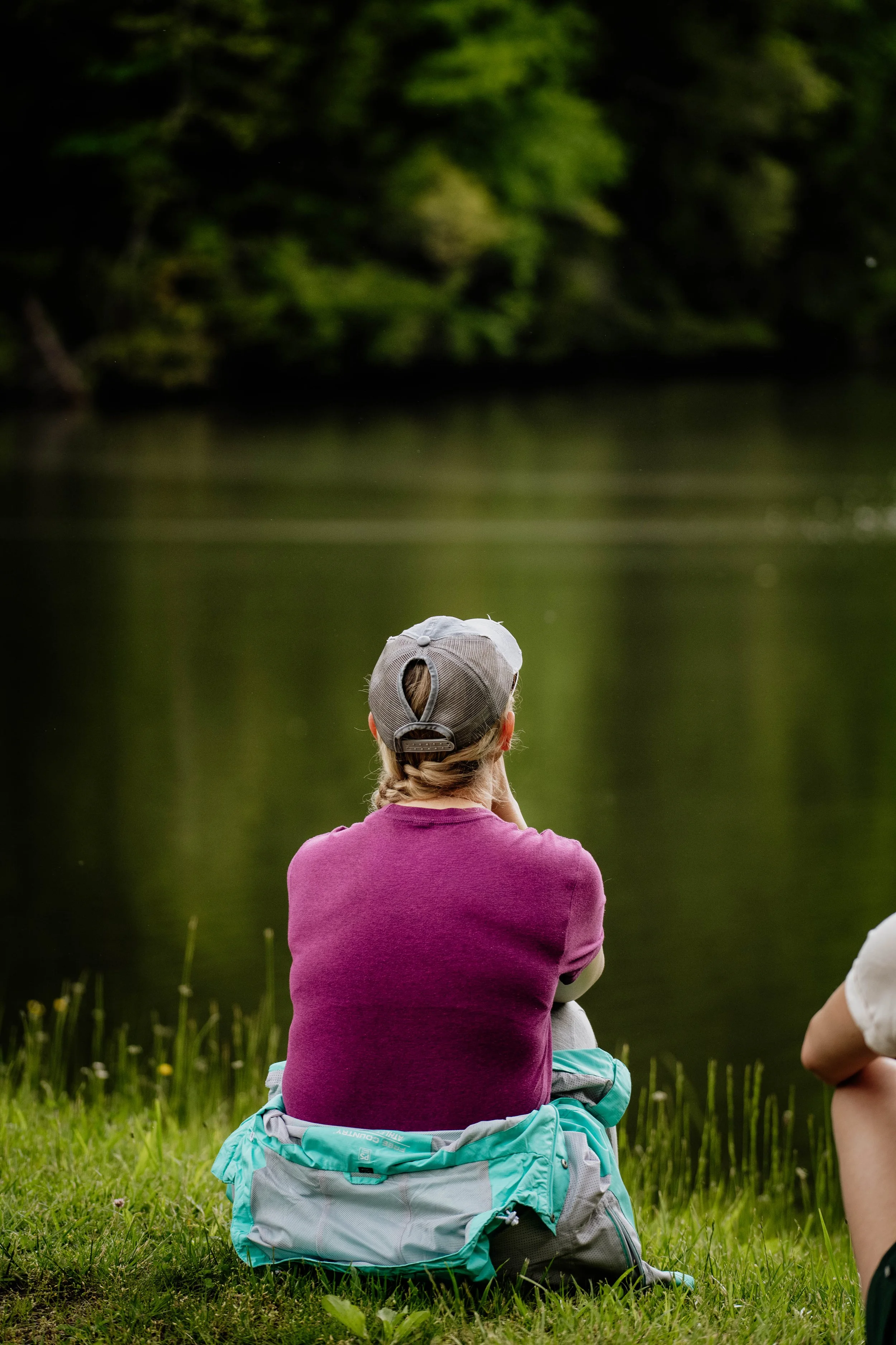 A person wearing a gray baseball cap and a purple shirt sitting by a lake, facing away, with green trees reflected in the water.