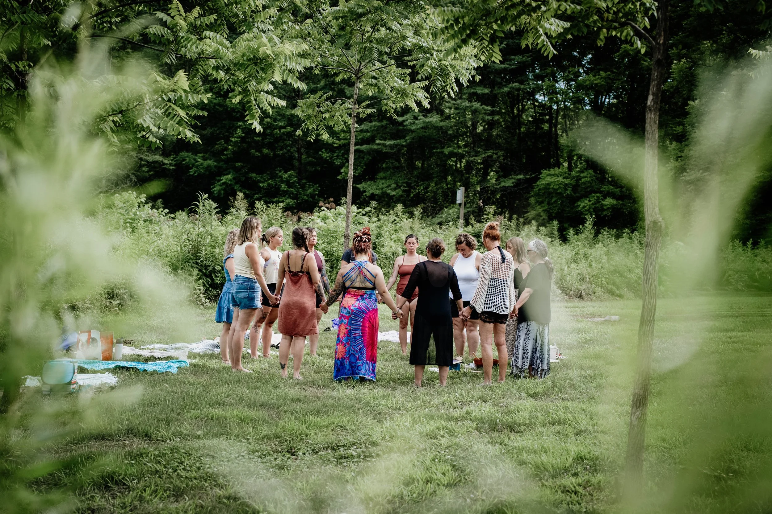 A group of people holding hands in a circle outdoors in a wooded area during the daytime.