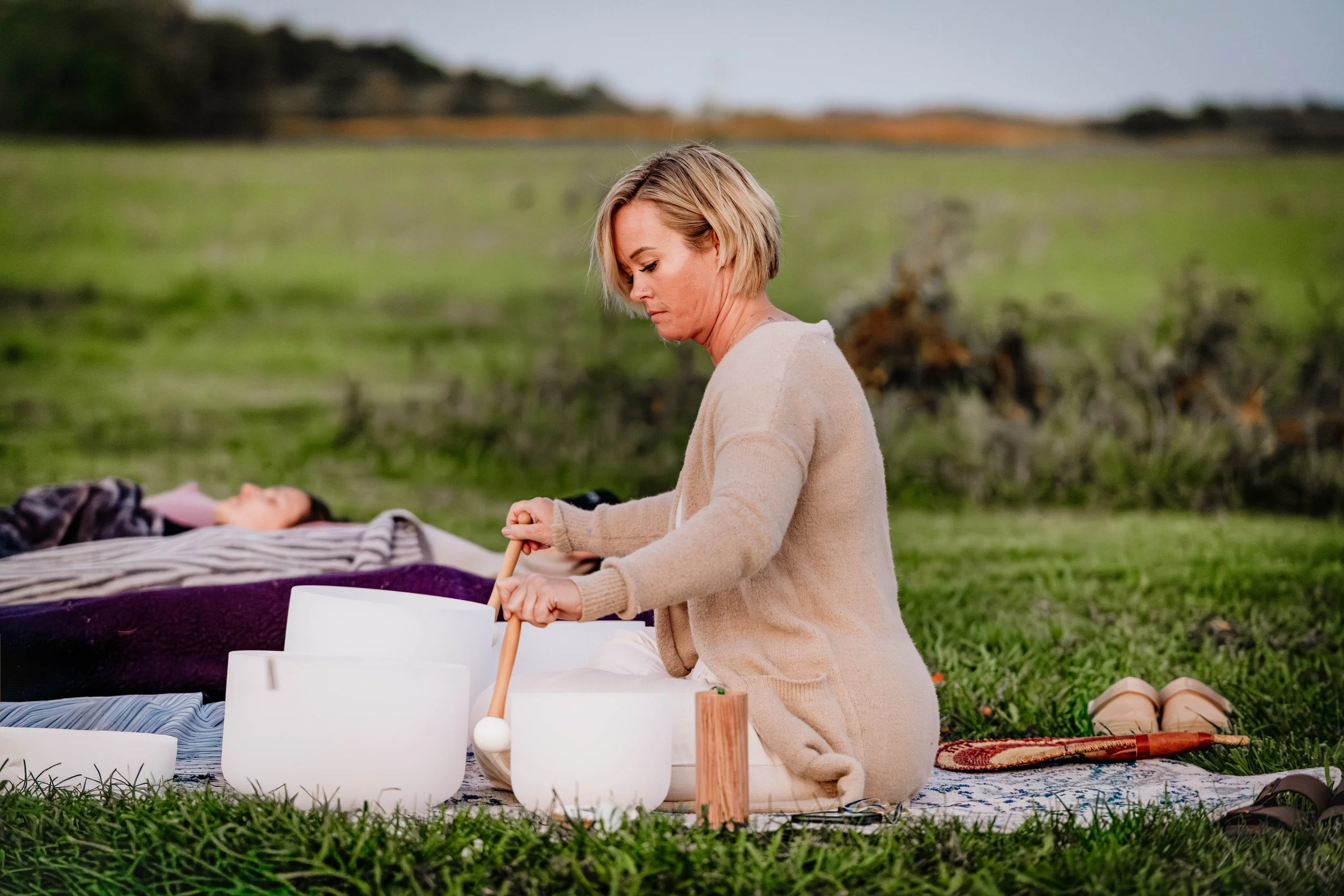 A woman kneels on a blanket outdoors, playing singing bowls, with a person lying down on the blanket. There are various objects around, including a wooden mallet and ceramic cups, in a grassy field with a blurred landscape in the background.