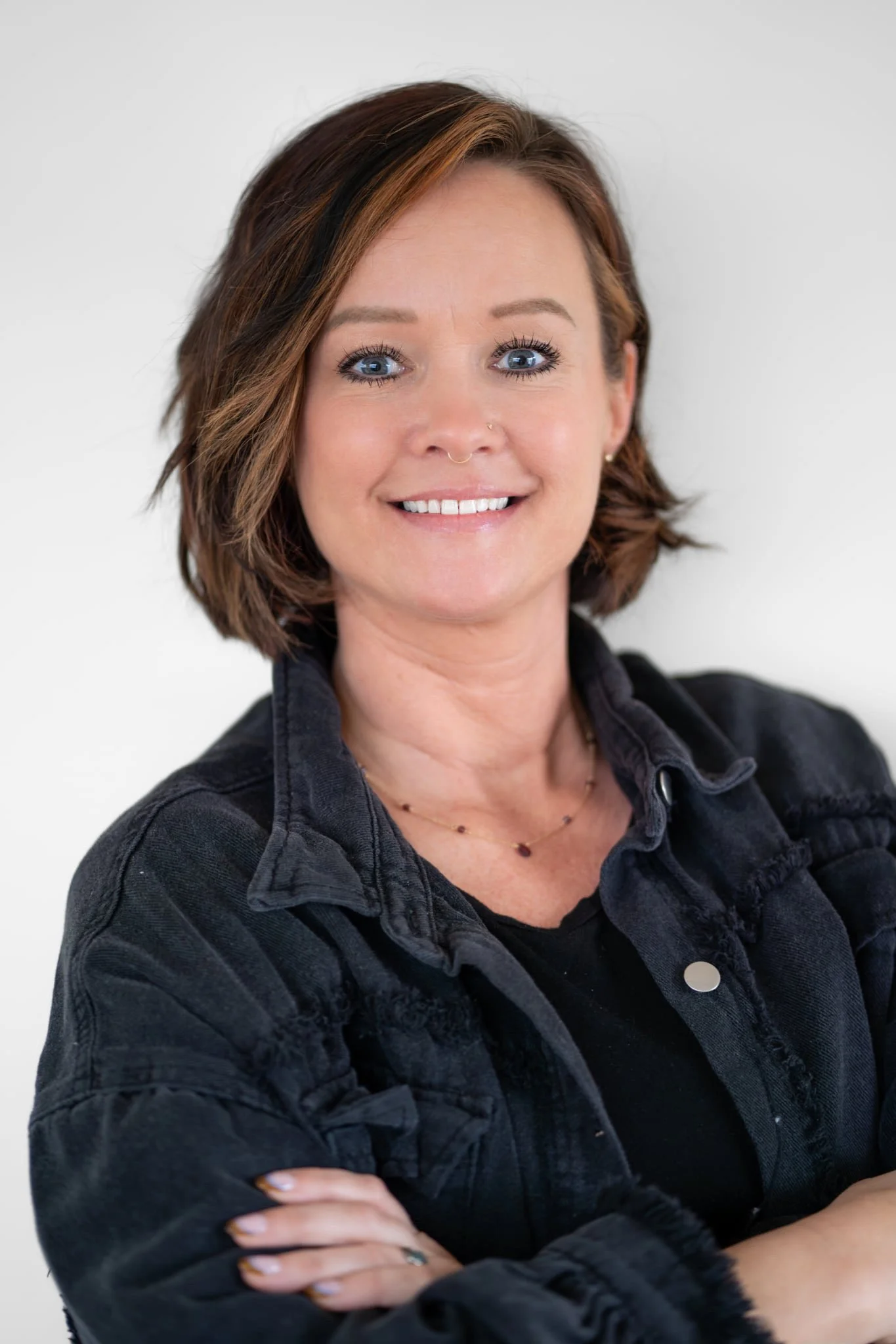 A woman with short brown hair and blue eyes, smiling, wearing a black shirt and a dark jacket, standing against a plain white background.