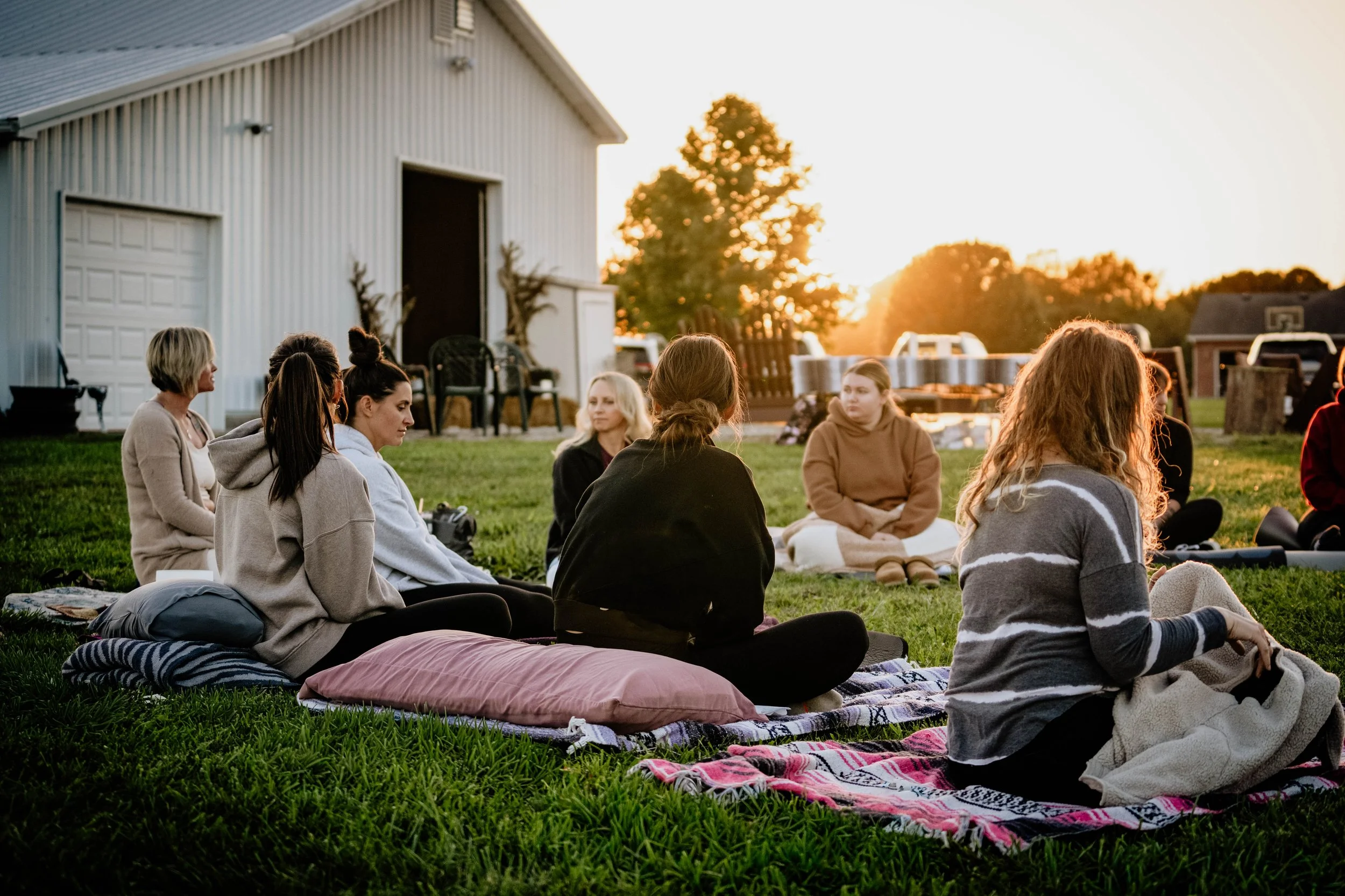 Group of people sitting on blankets outdoors in front of a large white building with garage doors, during sunset.
