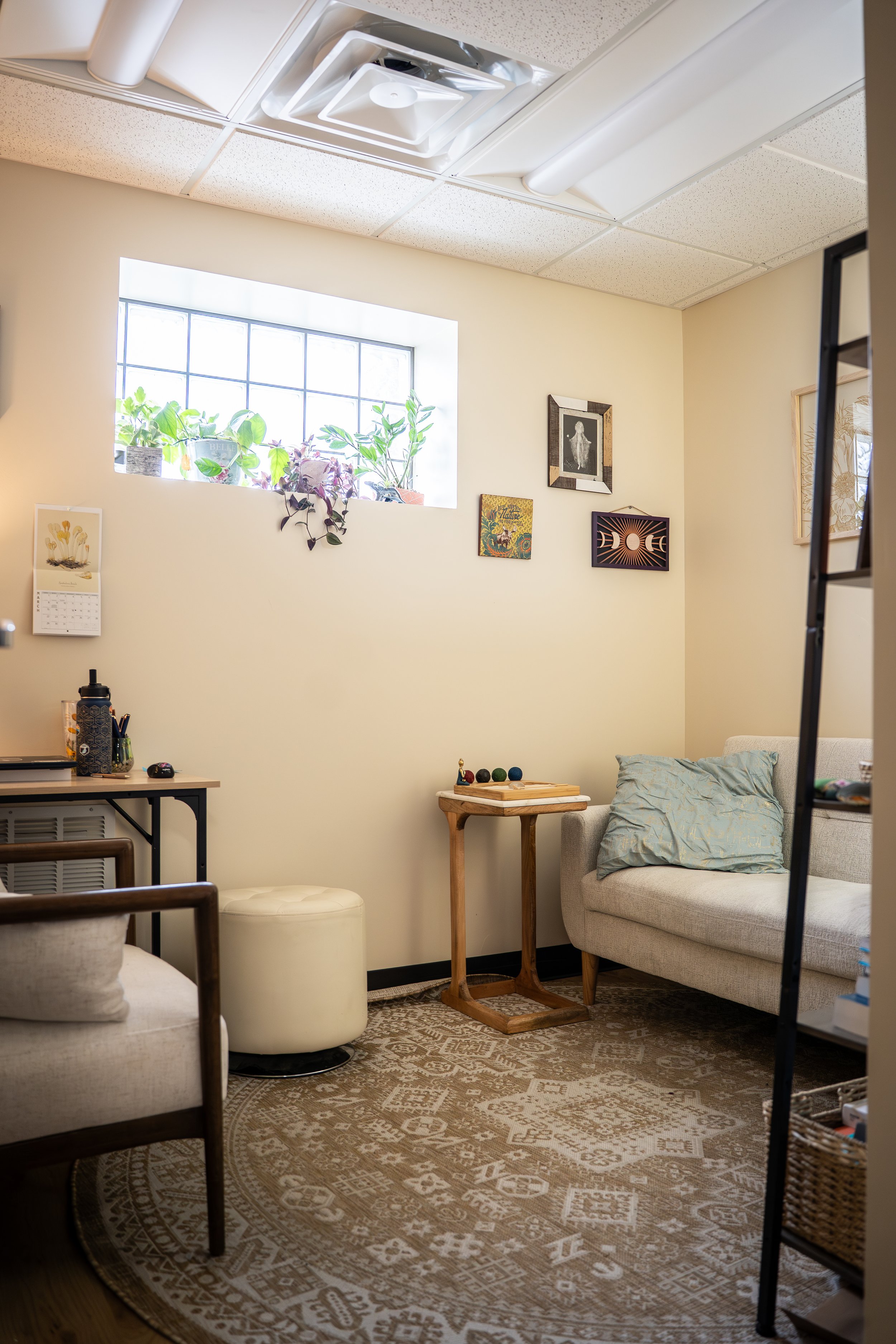 A cozy living room with a window, framed art on the wall, and furniture including a beige couch with a pillow, a small side table, and a patterned rug.